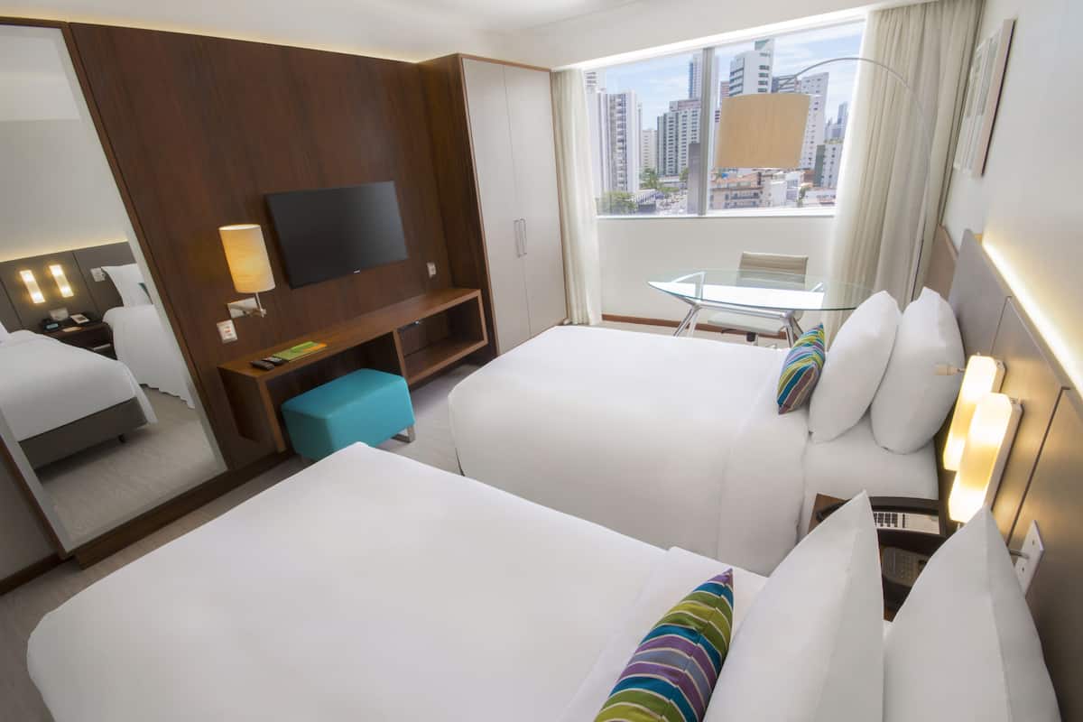 A hotel room with two beds, a TV, a desk, and a window with a city view.