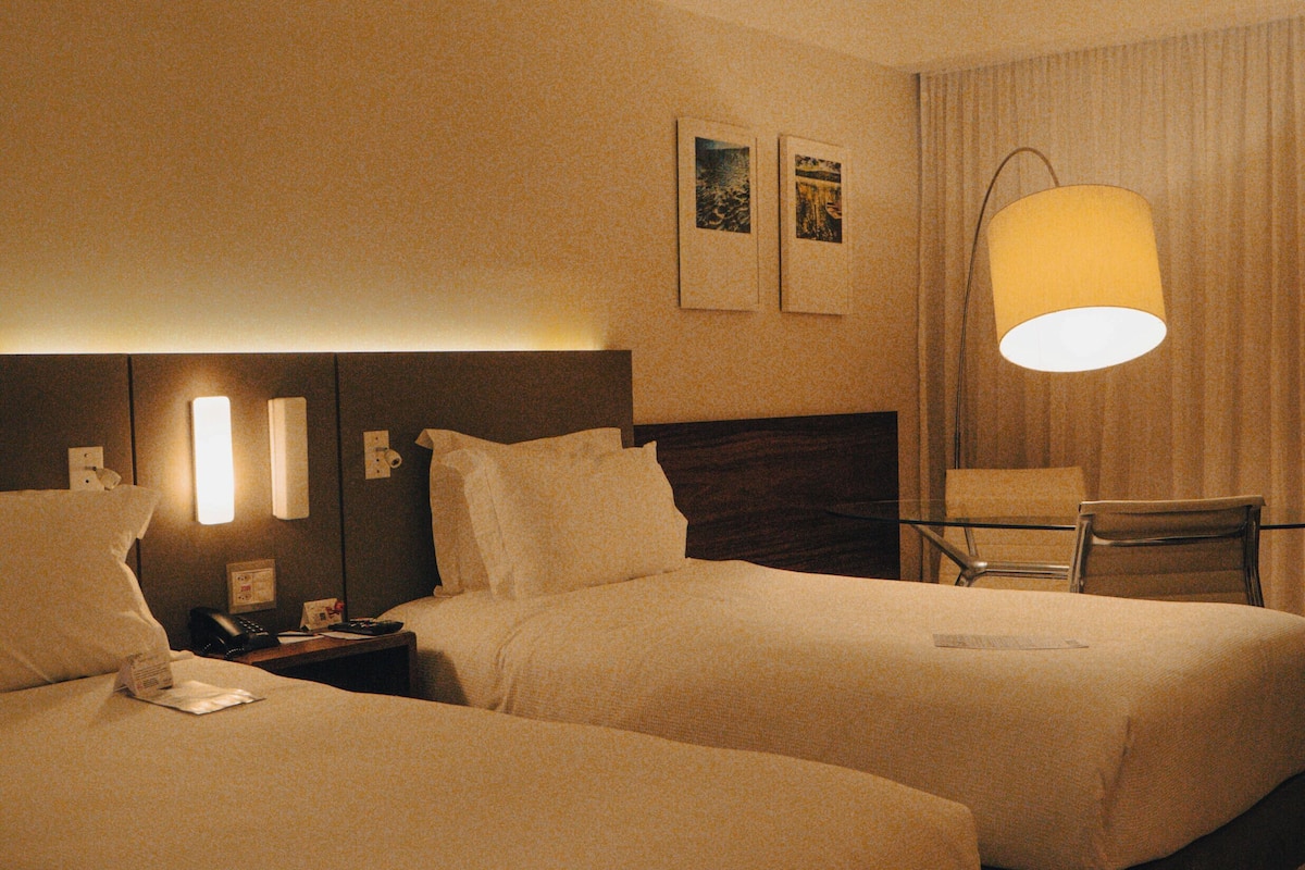 A hotel room with two beds, a nightstand with a lamp, a chair, and a wall-mounted telephone.