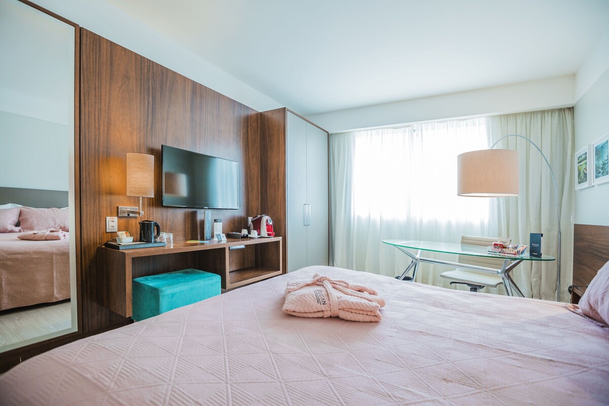 Habitación de hotel con cama, escritorio con lámpara, televisión y ventana con cortinas.