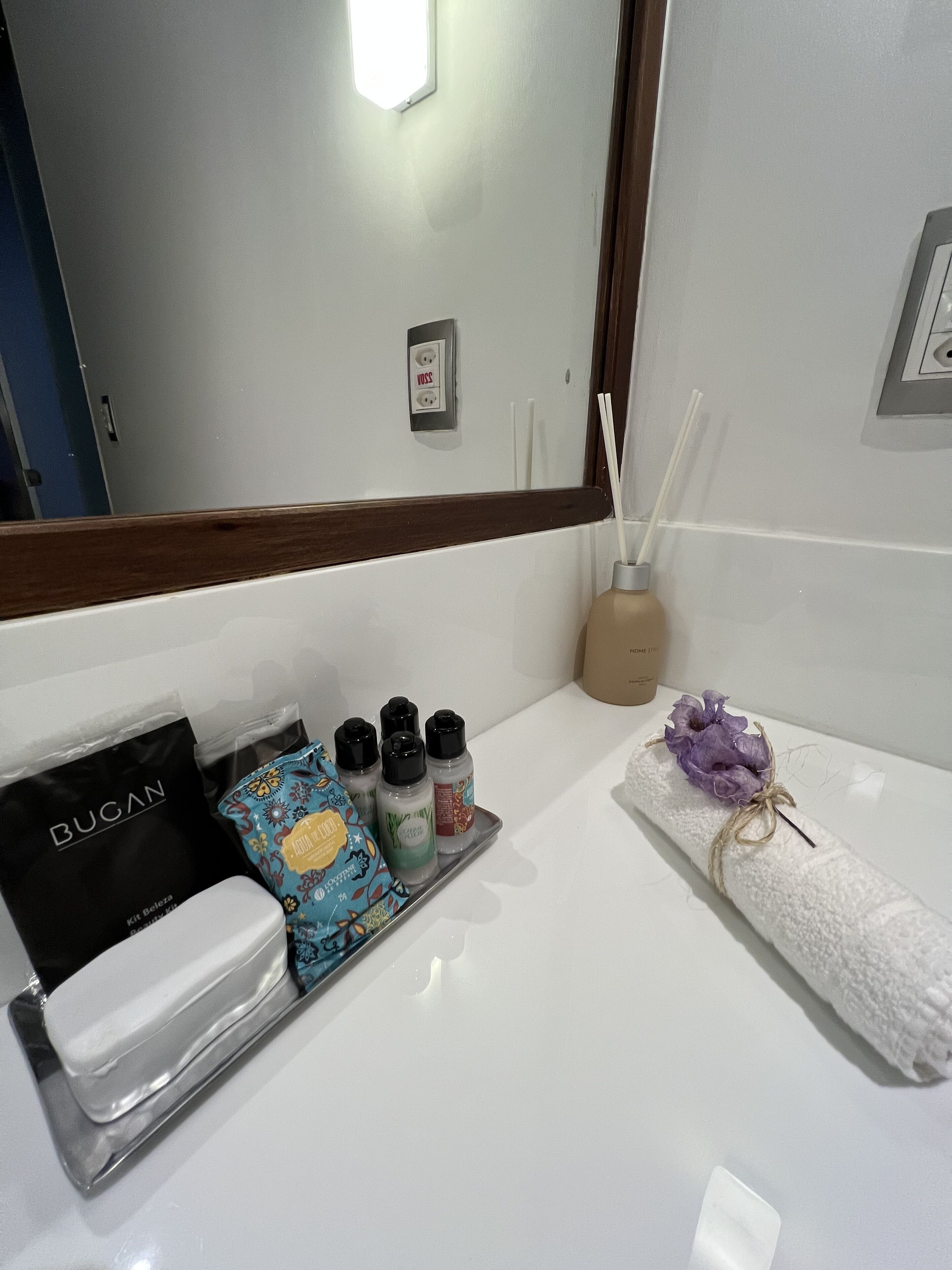 A bathroom with a white sink, a mirror, and toiletries including a towel, a soap dispenser, and a tray with a bag of Bugan.