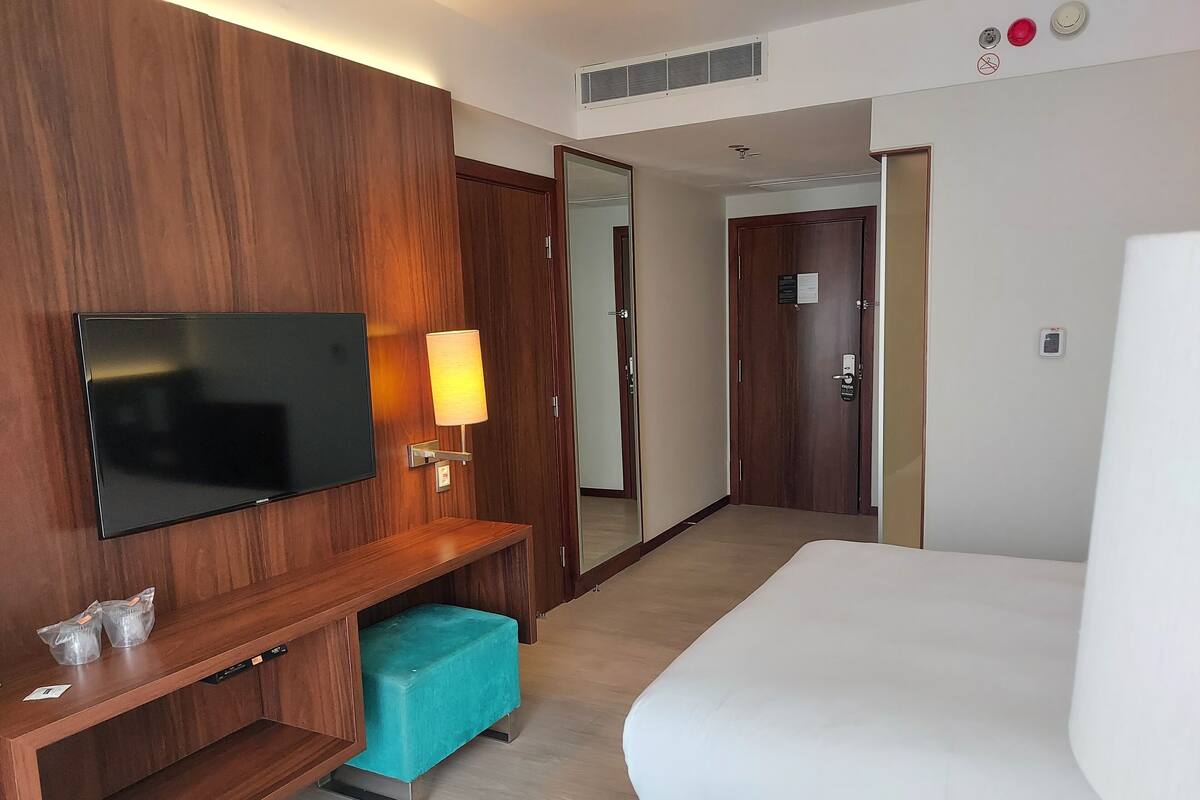 A hotel room with a flat-screen TV mounted on the wall, a bedside table with a lamp, a teal bench, and a large bed.