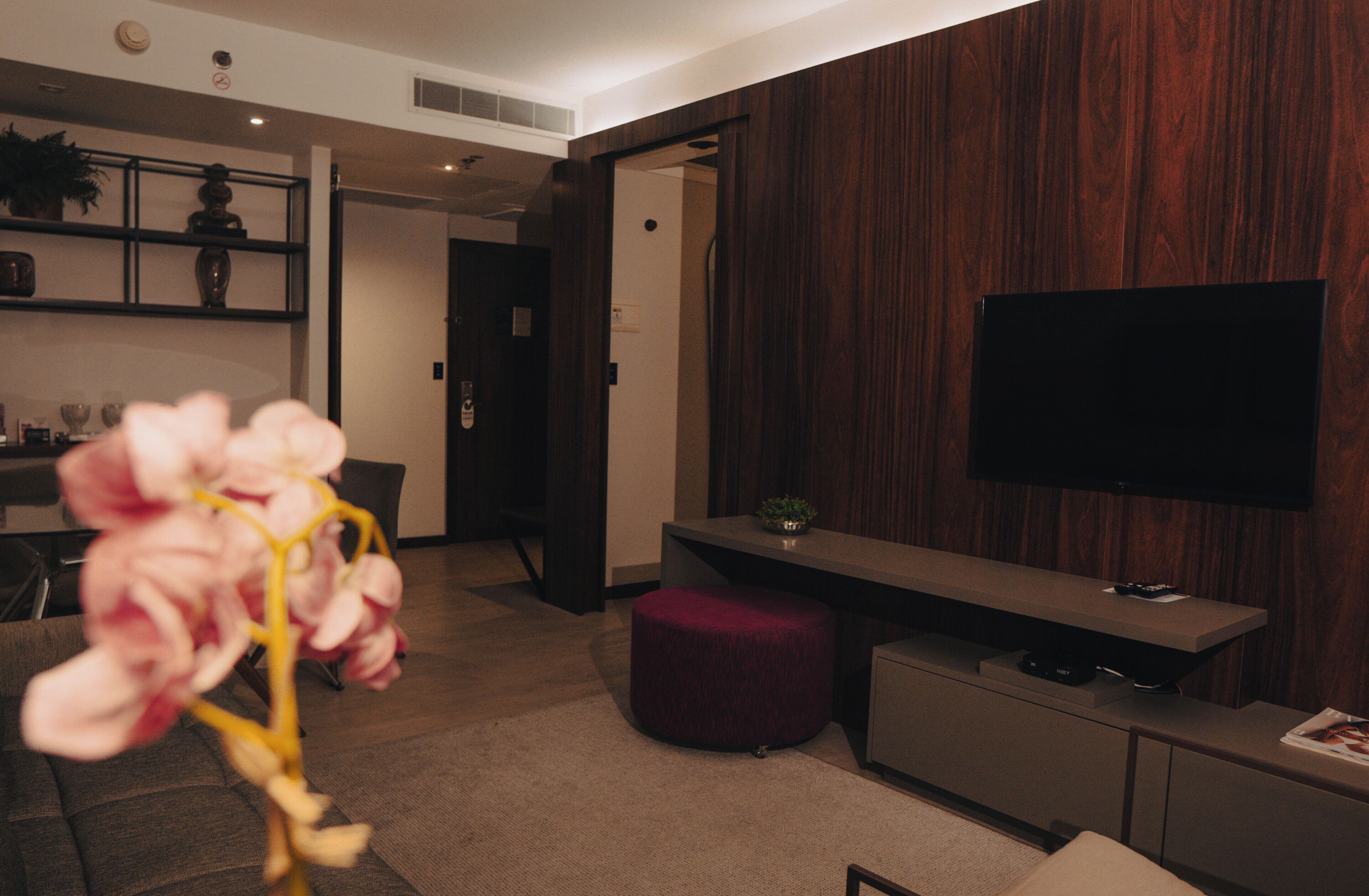 Un salon moderne avec un téléviseur à écran plat, un mur d’accent en bois foncé, un petit pouf rond et un coin repas à l’arrière-plan.
