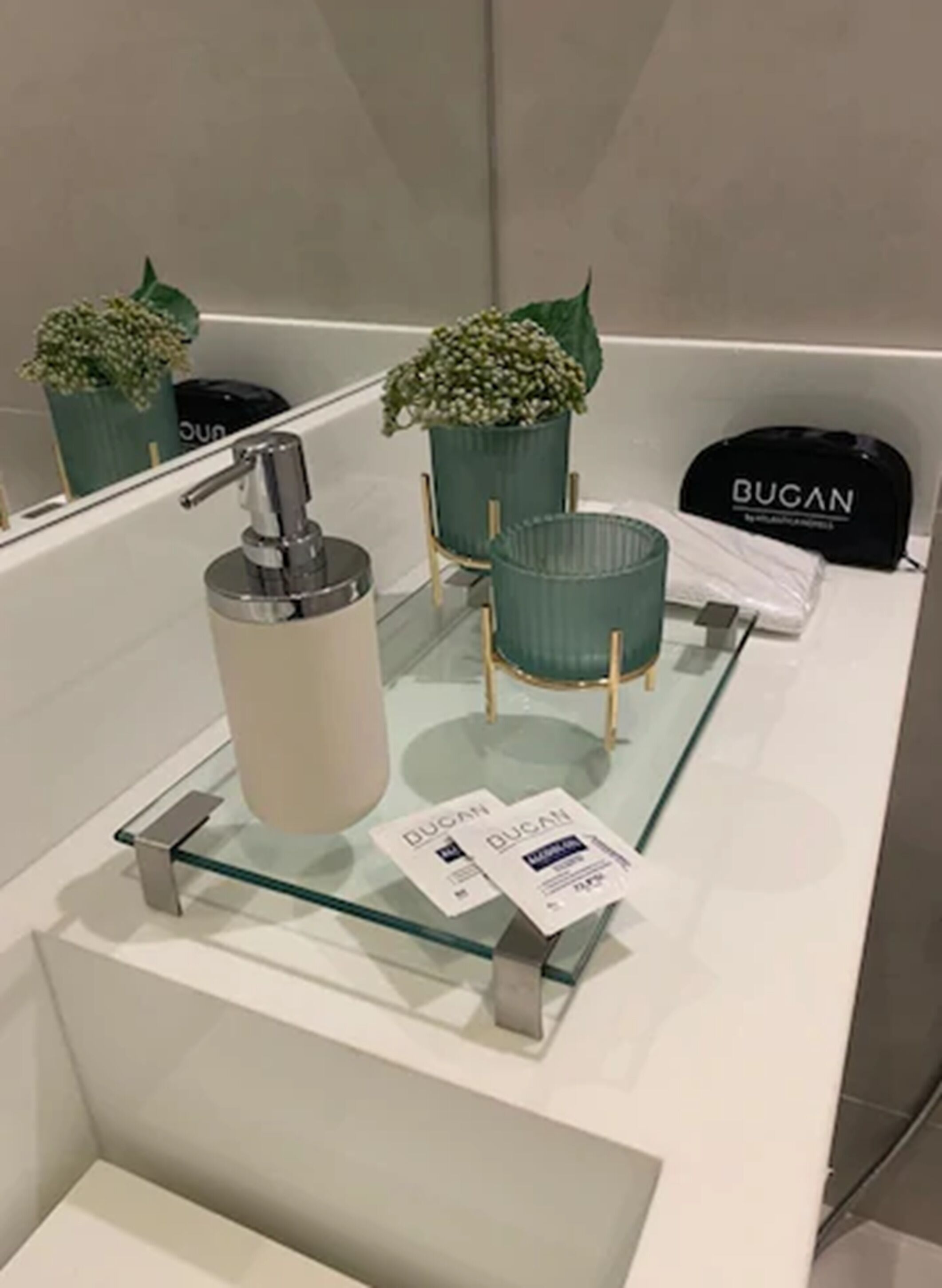 Un baño con un estante de vidrio que muestra una botella, una planta en maceta y una bolsa con el texto 'BUGAN'.