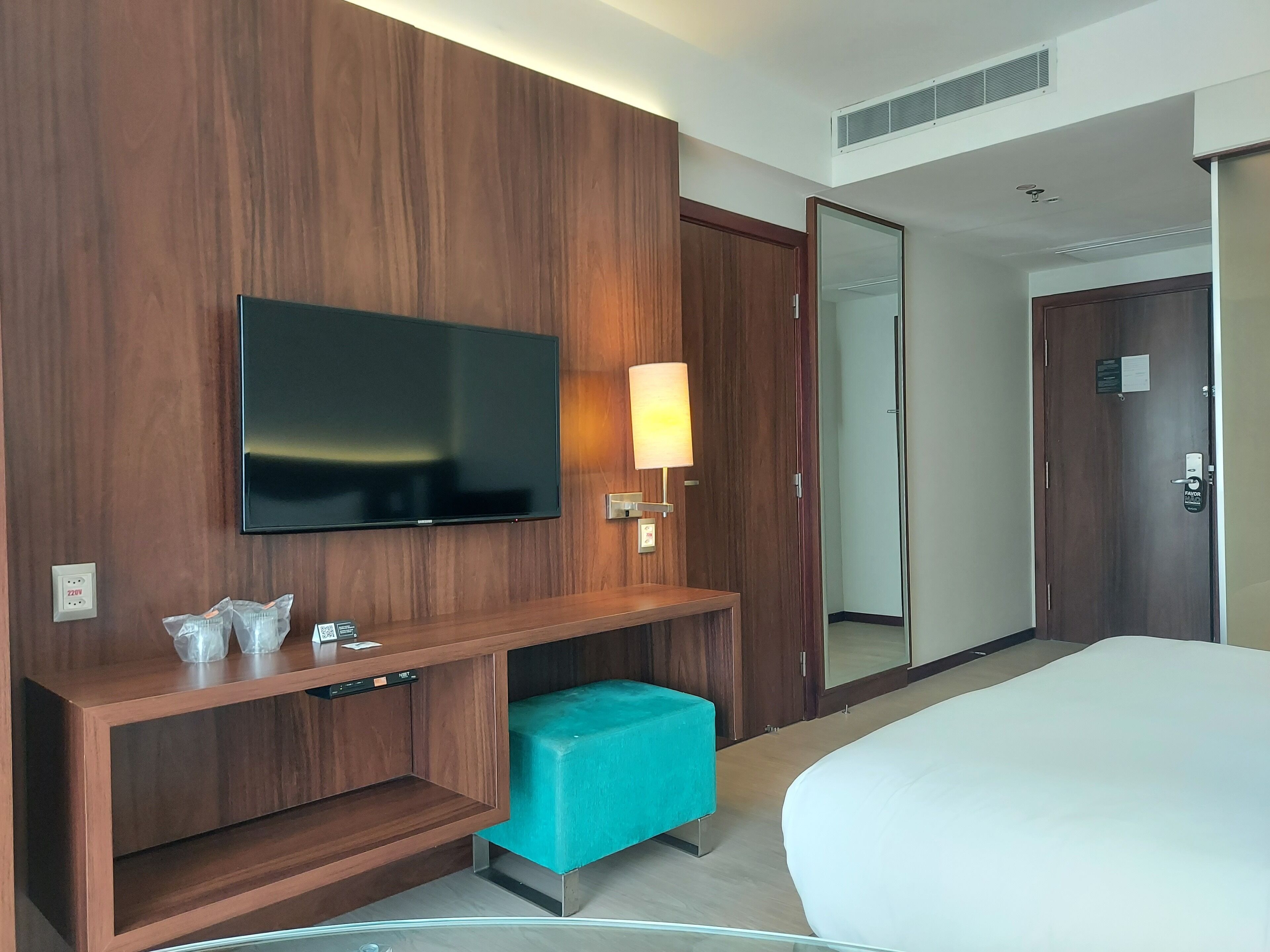 A hotel room with a large flat-screen TV mounted on a wooden wall, a bedside table with a lamp, a small teal bench, and a large bed.