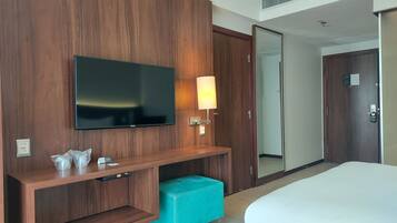 A hotel room with a large flat-screen TV mounted on a wooden wall, a bedside table with a lamp, a small teal bench, and a large bed.