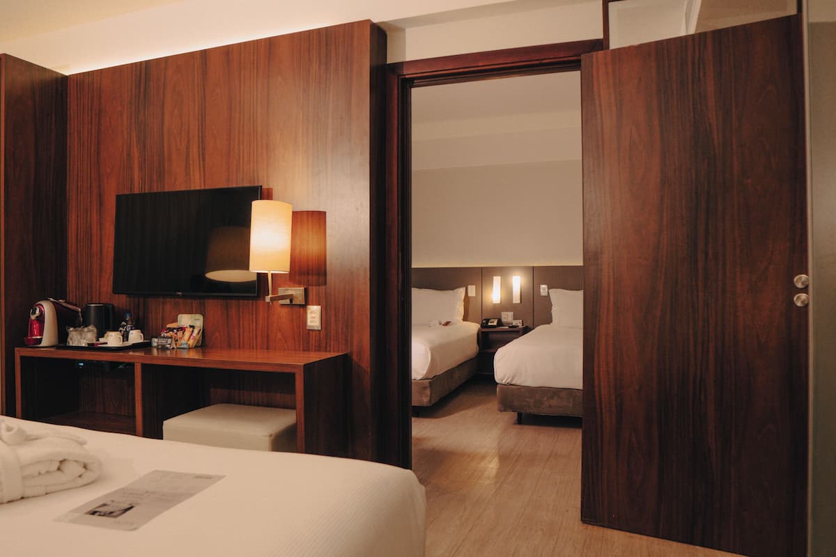 A hotel room with a wooden wall panel, a flat-screen TV, a desk with a lamp, a bed, and a bedside table.
