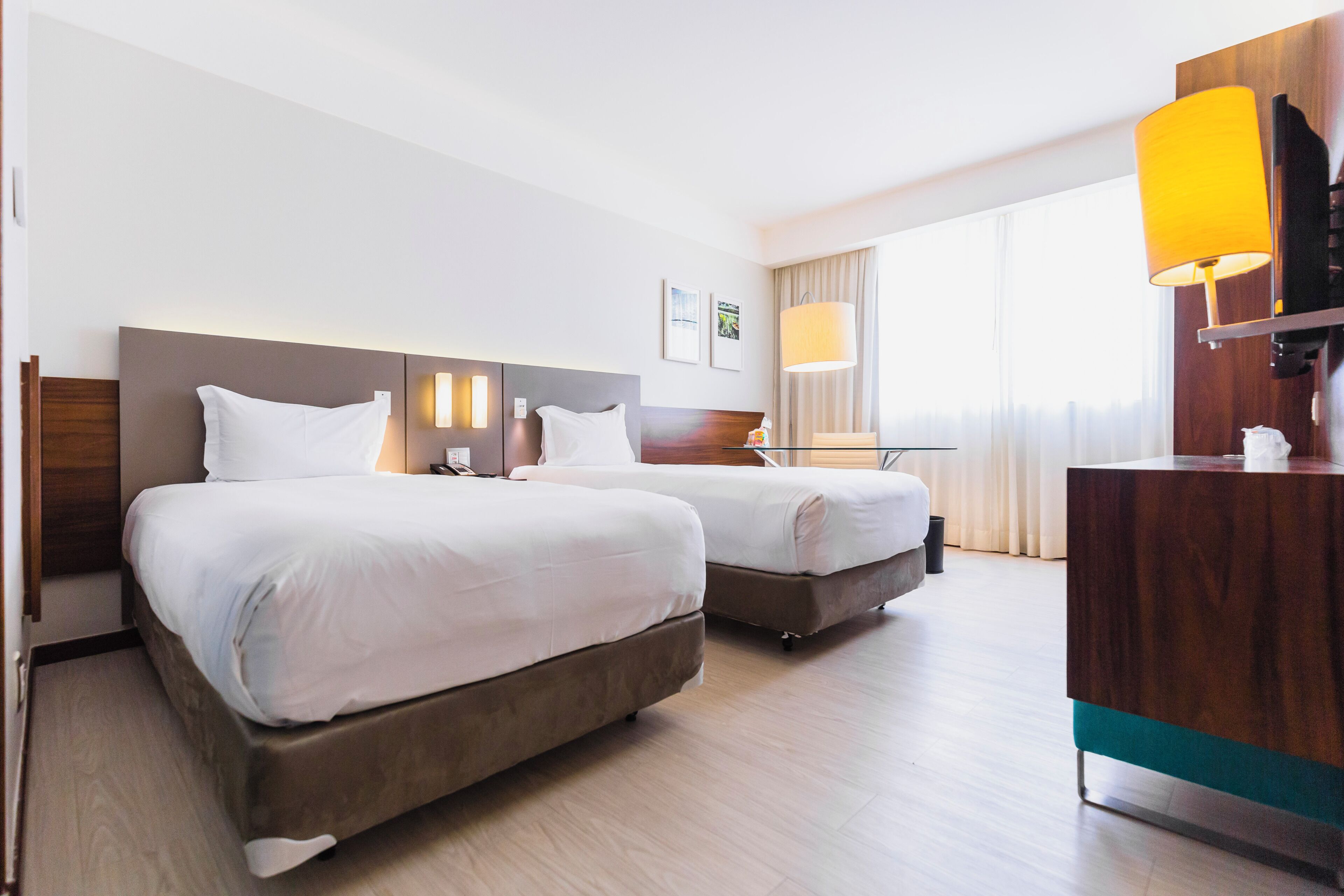 A hotel room with two beds, a wooden headboard, a desk with a lamp, and a large window with curtains.