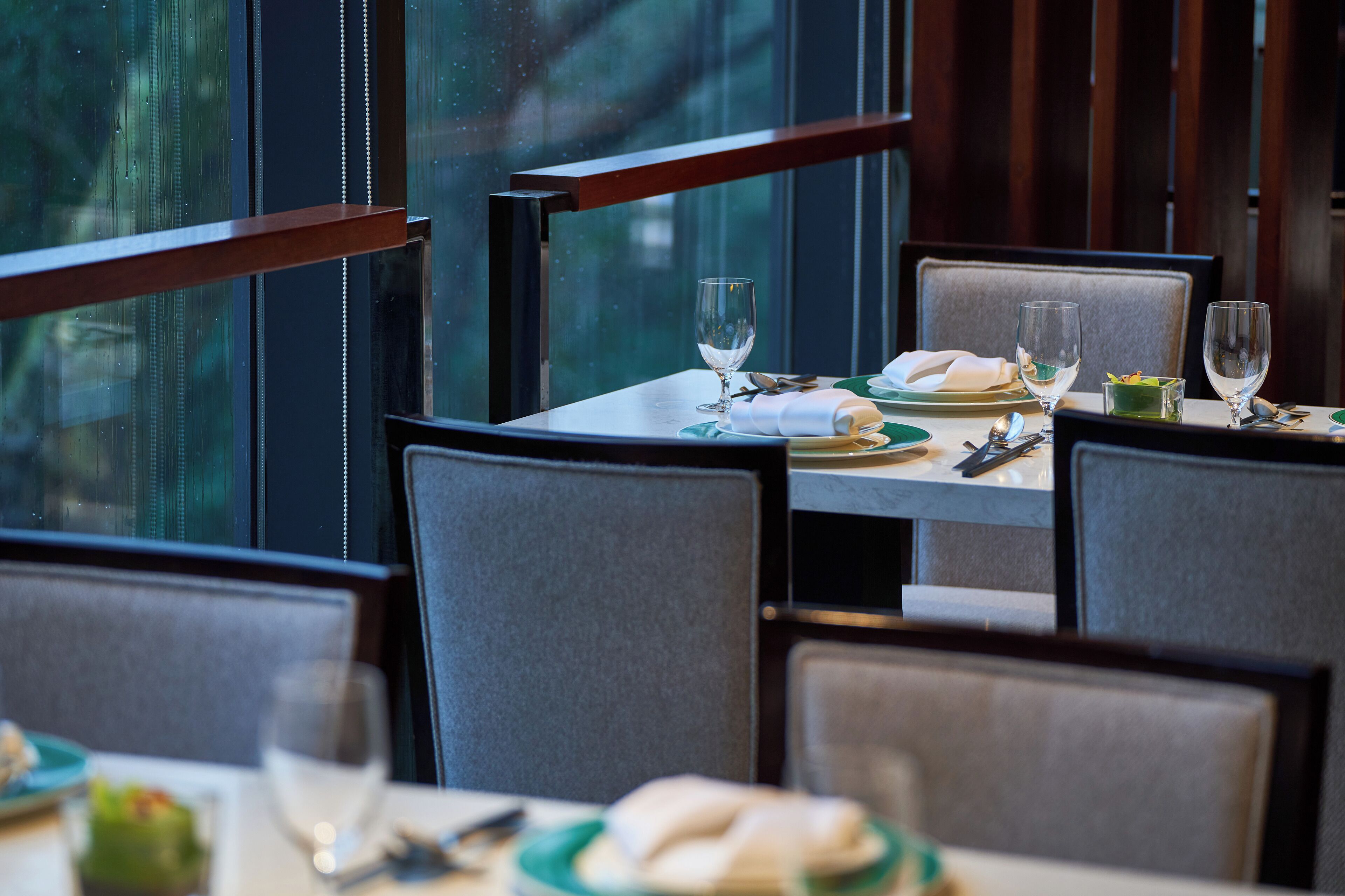 Un comedor con una mesa dispuesta para dos, sillas y vistas al exterior a través de amplios ventanales.