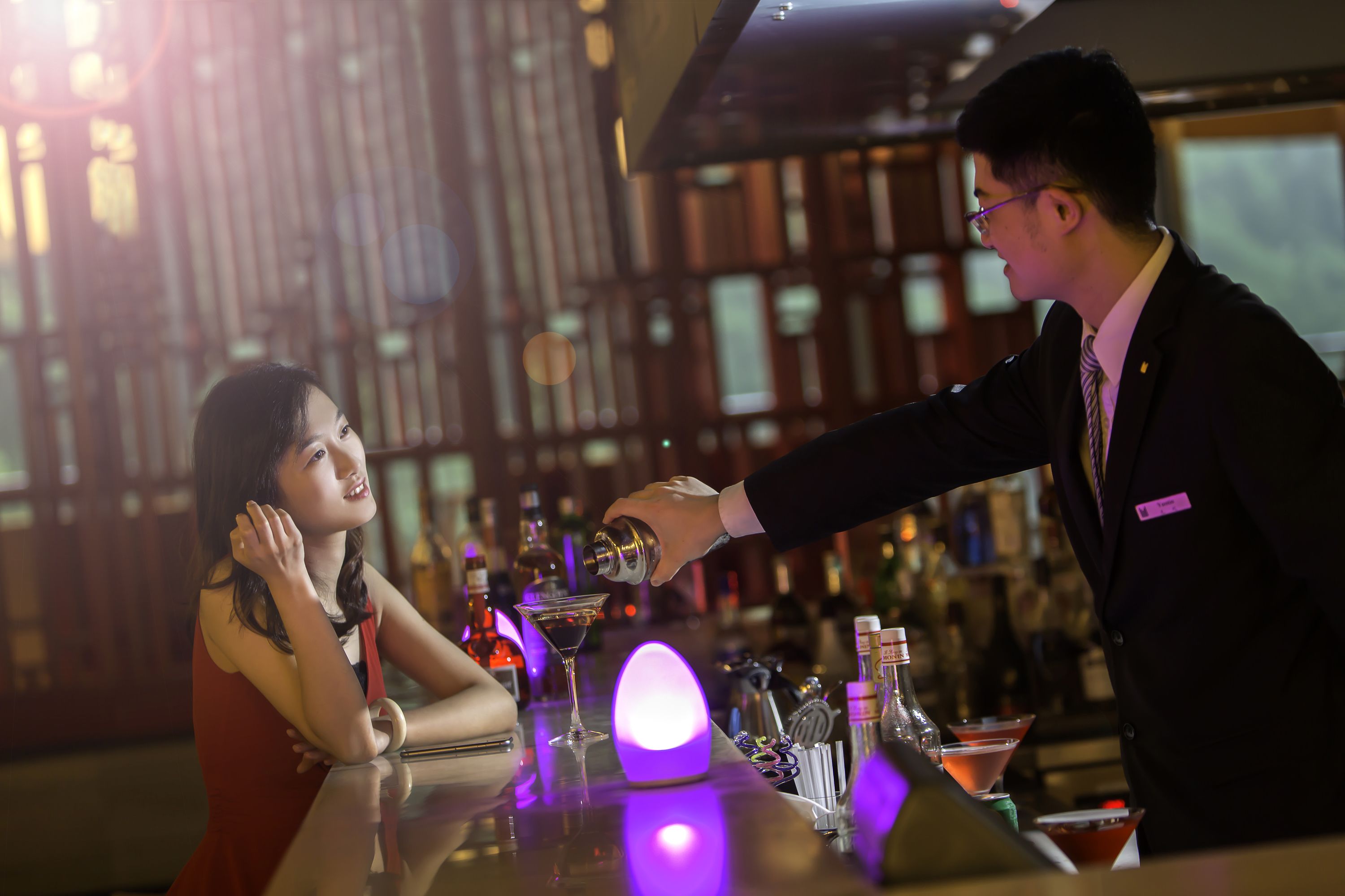 Une scène de bar : on voit une personne en train de verser une boisson dans un verre à martini, une femme assise au bar, ainsi que diverses bouteilles et différents verres sur le comptoir.