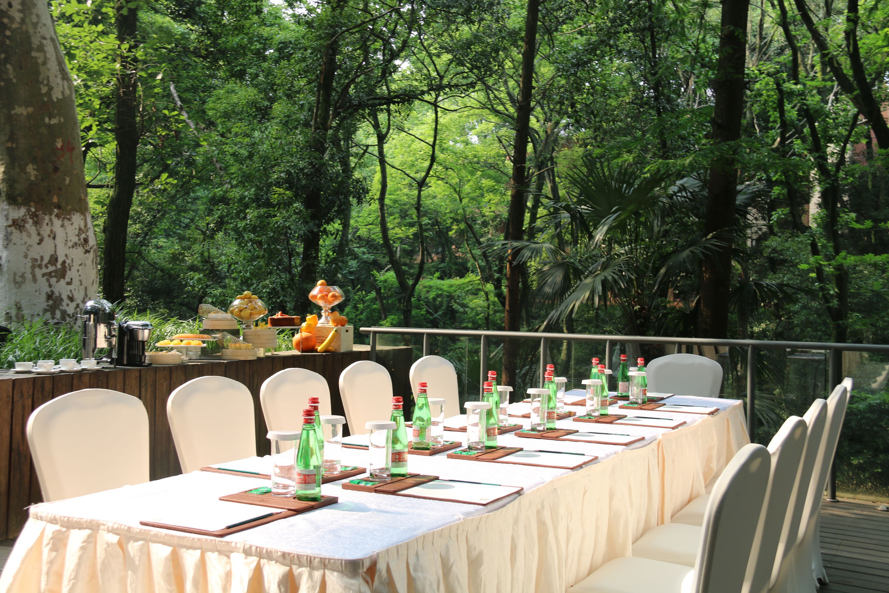 A long dining table set outdoors with white chairs, surrounded by lush greenery.