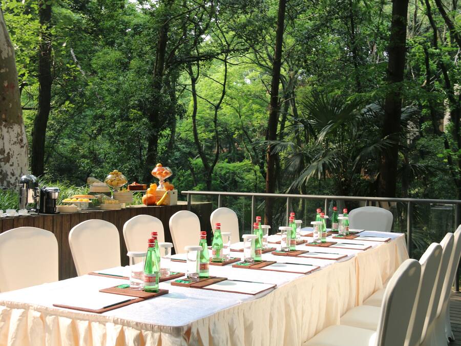 A long dining table set outdoors with white chairs, surrounded by lush greenery.
