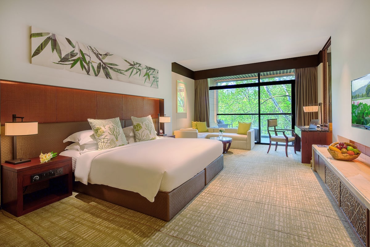 Une chambre d’hôtel avec un grand lit, un bureau, une chaise et une vue sur la verdure par la fenêtre.
