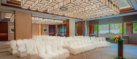 A spacious hall with rows of white chairs, a modern ceiling design, and large windows with greenery outside.