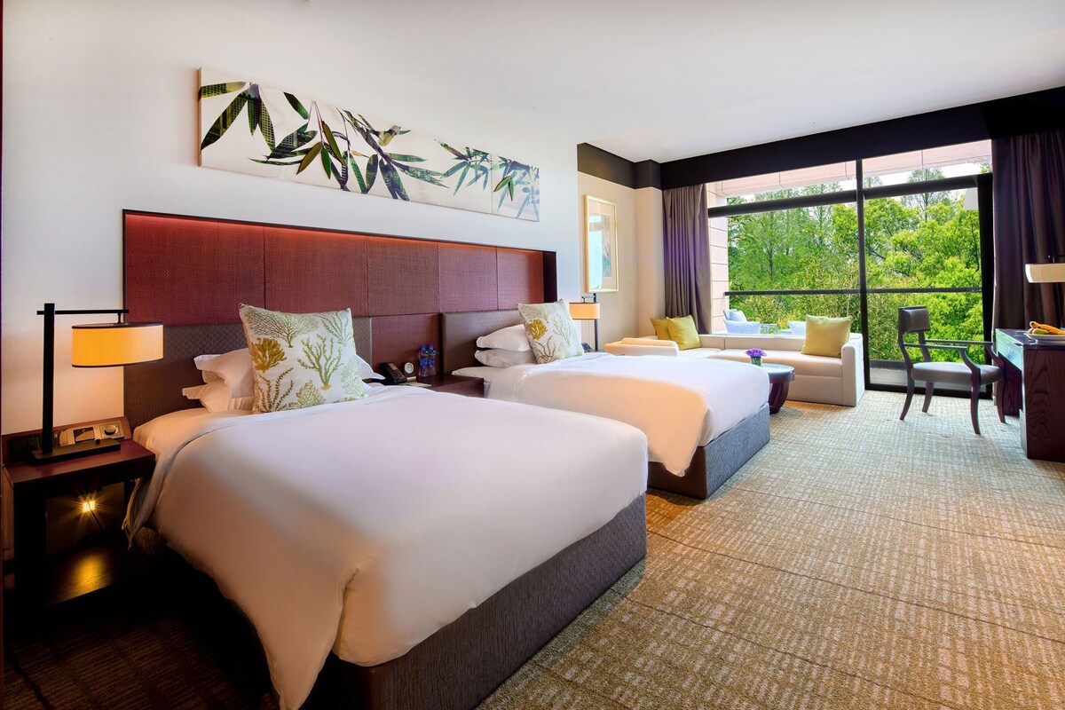 Quarto de hotel com duas camas, um sofá, uma escrivaninha e uma janela grande com vista para o verde.
