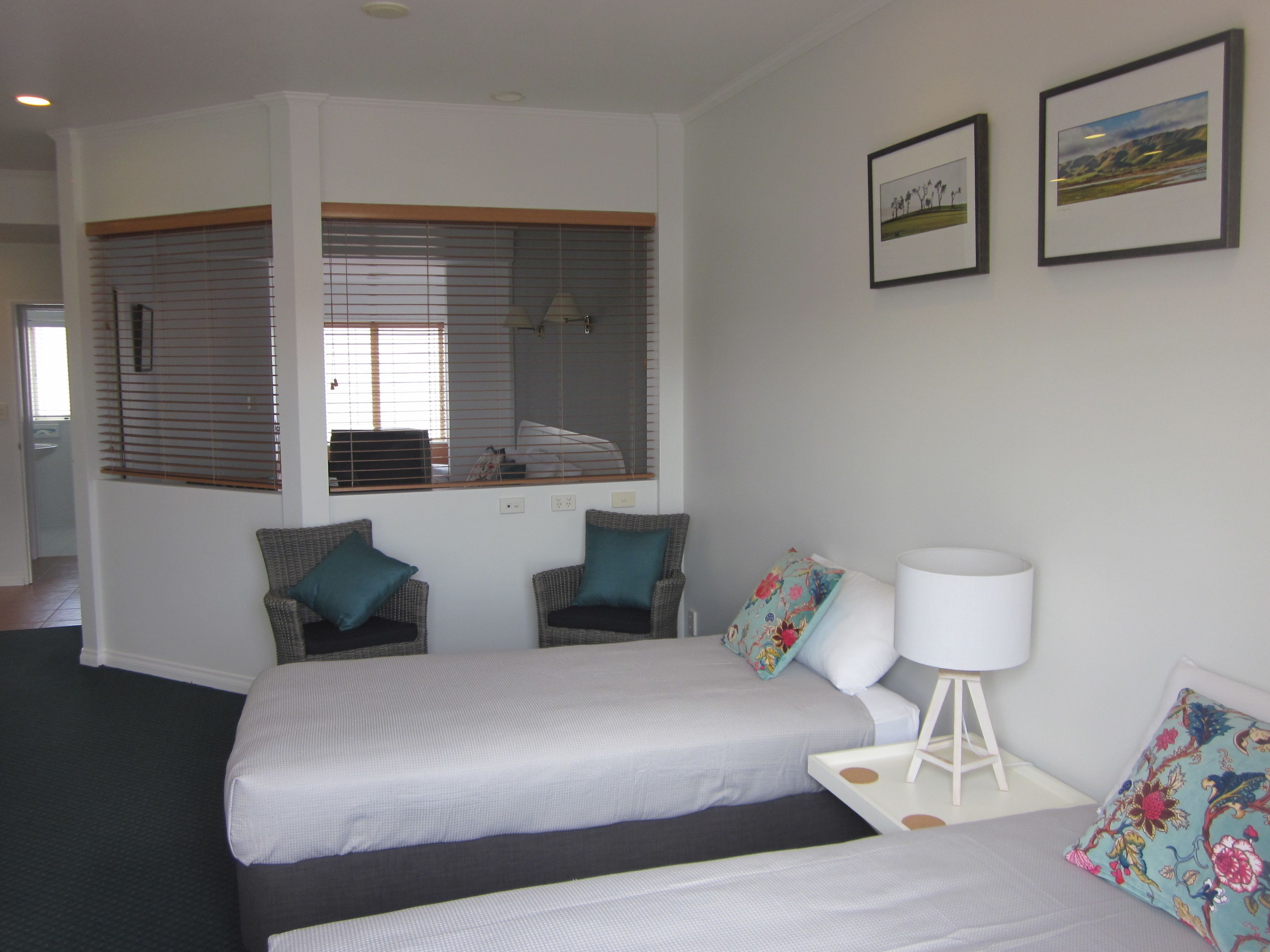 A hotel room with two beds, a large window with blinds, a small table with a lamp, and two framed pictures on the wall.