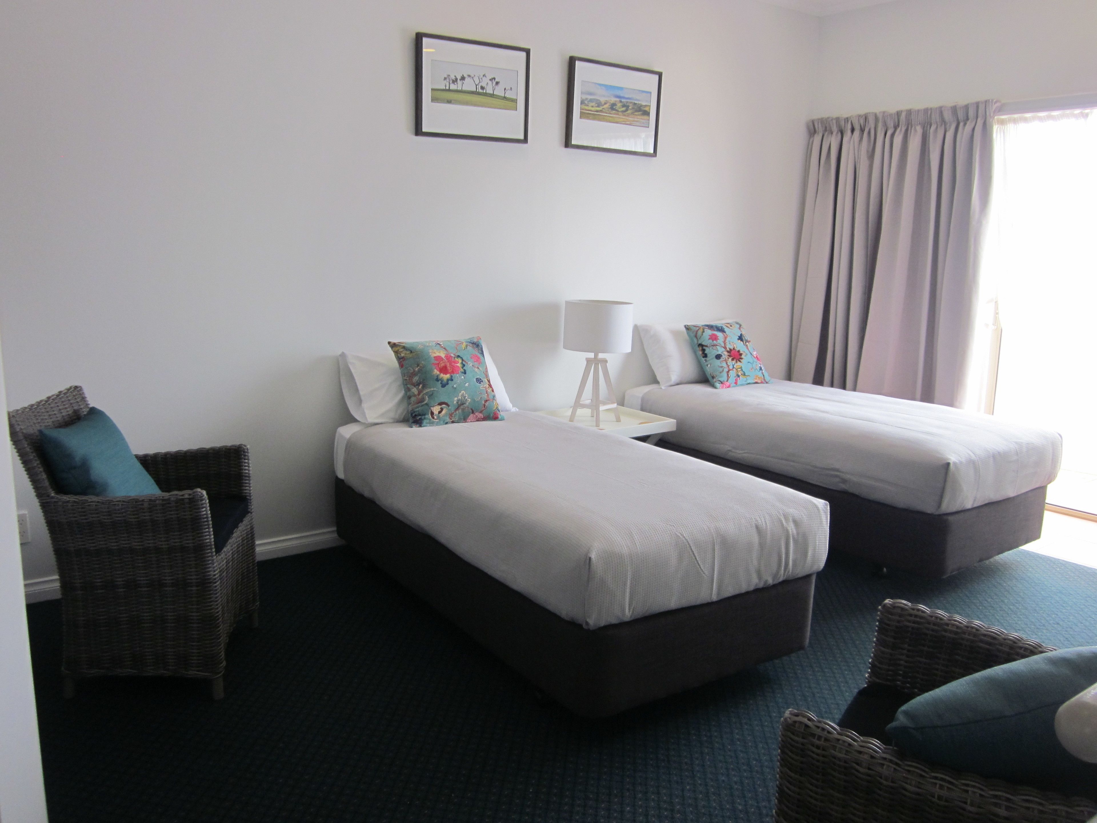 A hotel room with two single beds, a chair, a small table with a lamp, and two framed pictures on the wall.