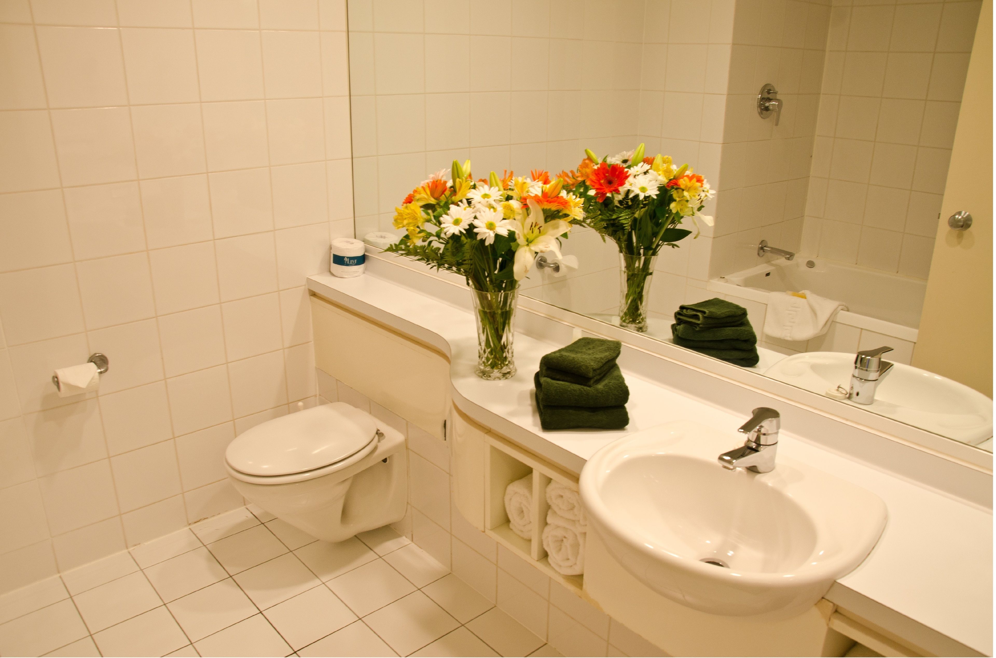 Ett badrum med toalett, handfat och spegel. Det finns handdukar och en vas med blommor på hyllan.