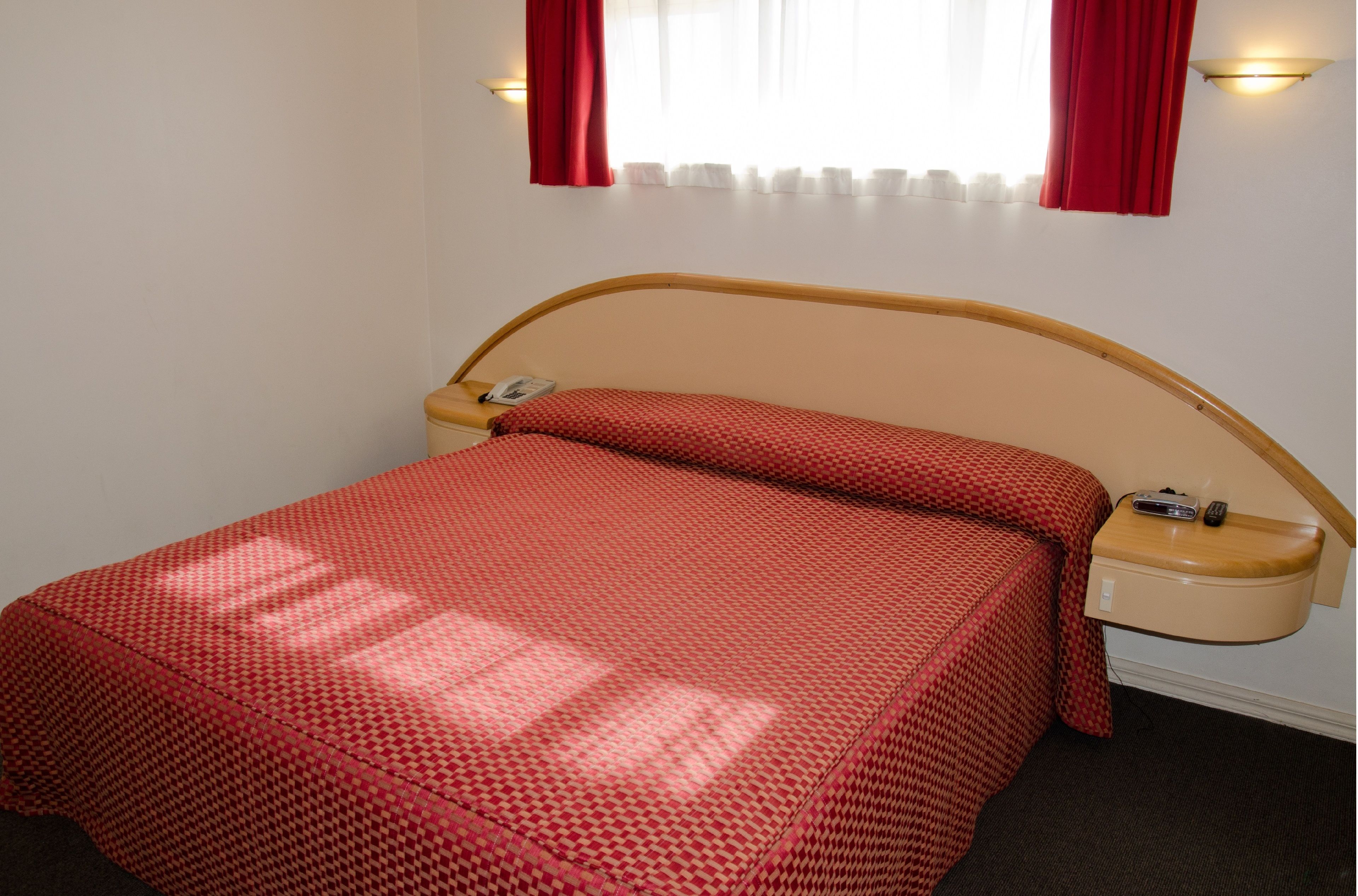 A single bed with a wooden headboard, a bedside table with a lamp, and a window with red curtains.
