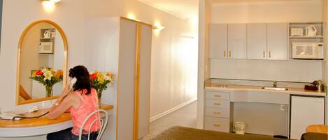 A compact hotel room with a bed, a desk with a telephone, a mirror, a small kitchenette with a microwave, and a wardrobe.