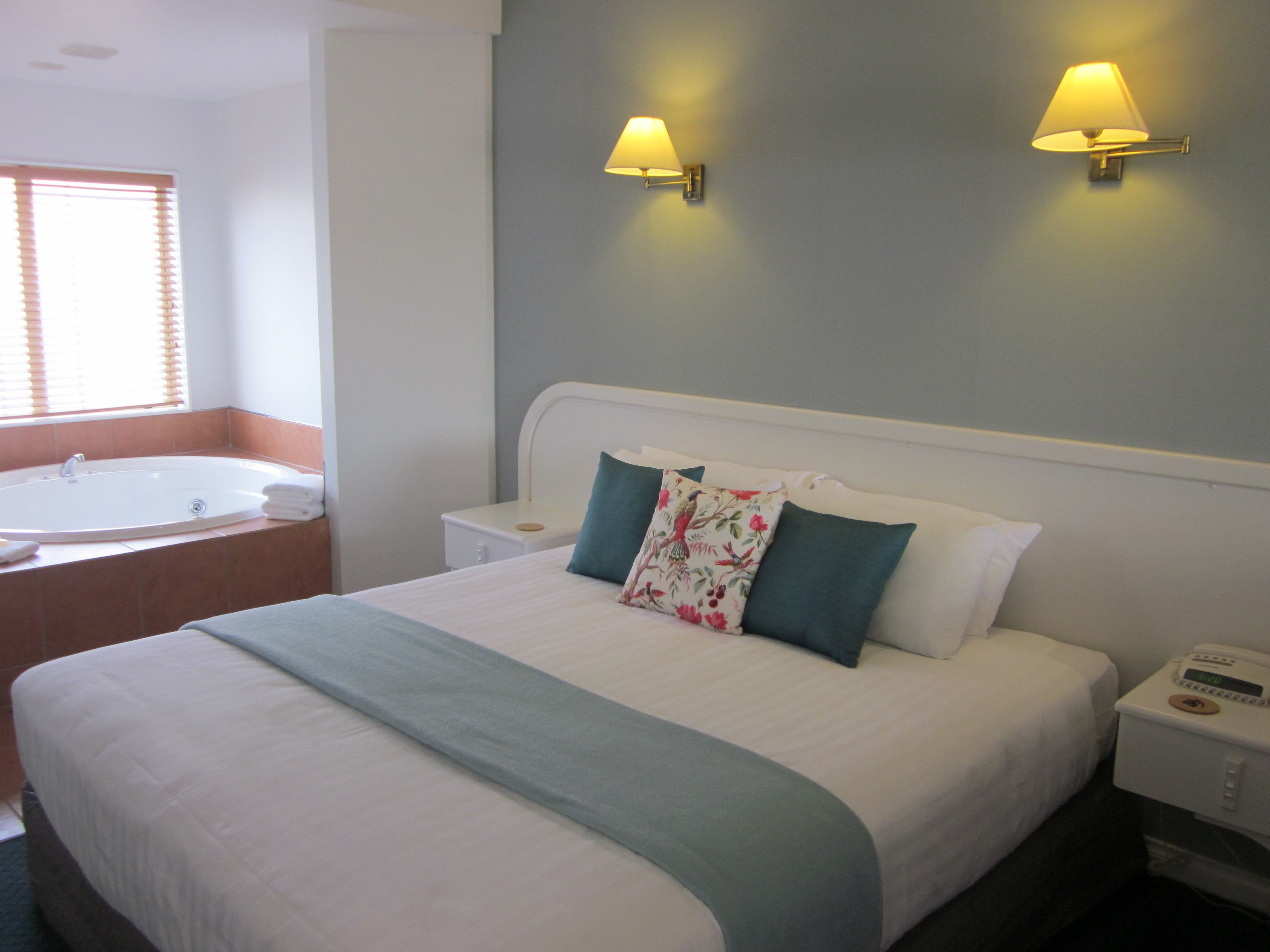 A hotel room with a large bed, a bedside table, a nightstand, a wall-mounted lamp, a window with blinds, and a bathroom with a bathtub.