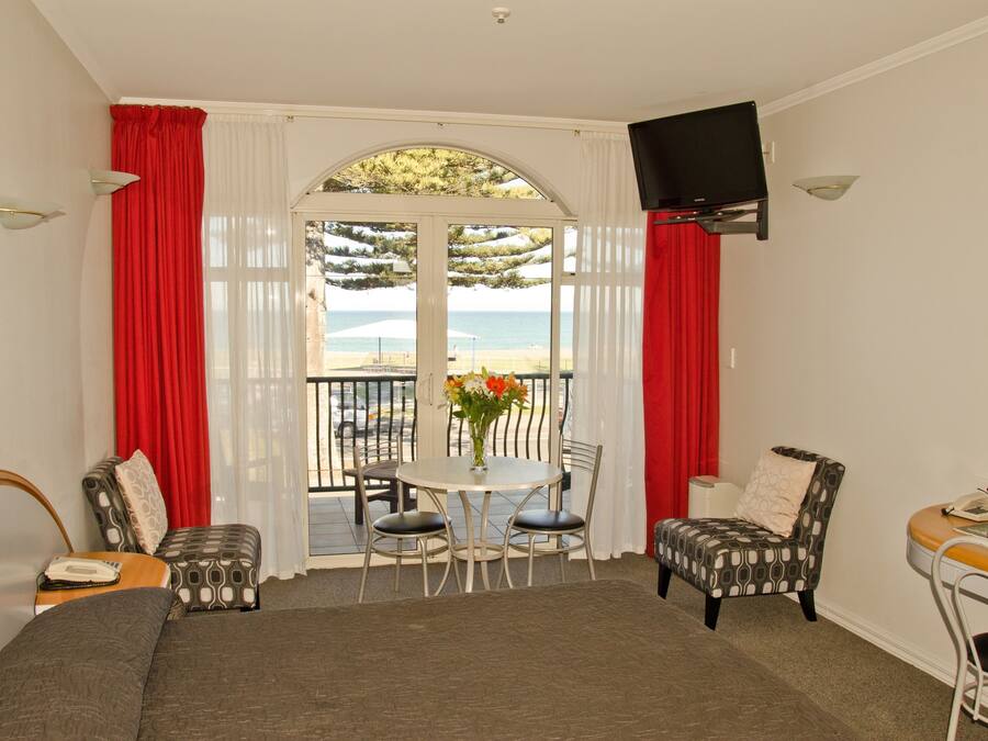 A room with a bed, a table with chairs, a TV mounted on the wall, and a view of the sea through a window.