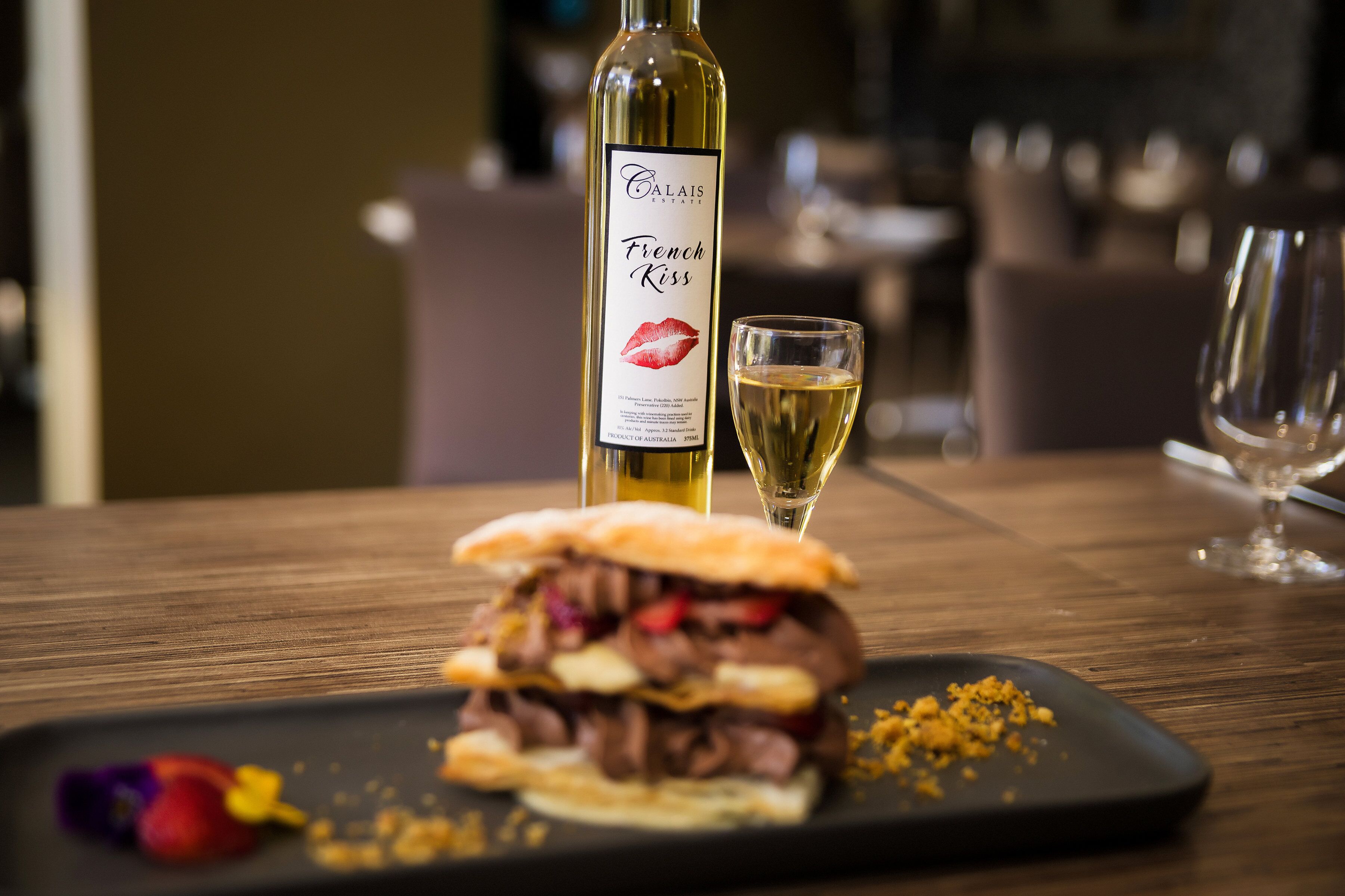 A sandwich on a black plate with a bottle of wine and a glass of wine on a wooden table.