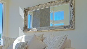 A living room with a large ornate mirror on the wall, a beige sofa with patterned cushions, and a window providing natural light.