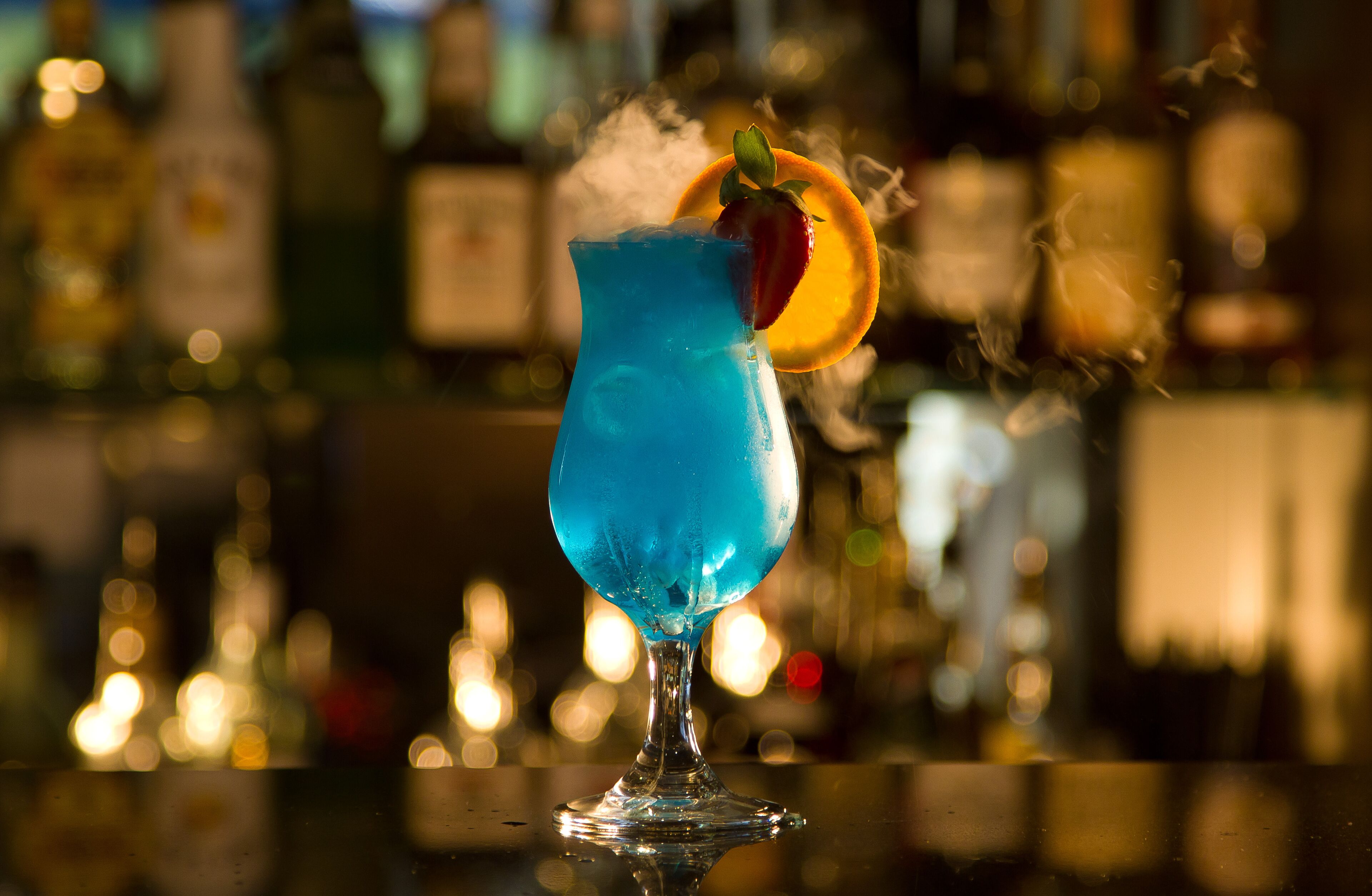 A cocktail in a glass with a strawberry garnish and an orange slice, with a blurred background of bottles.