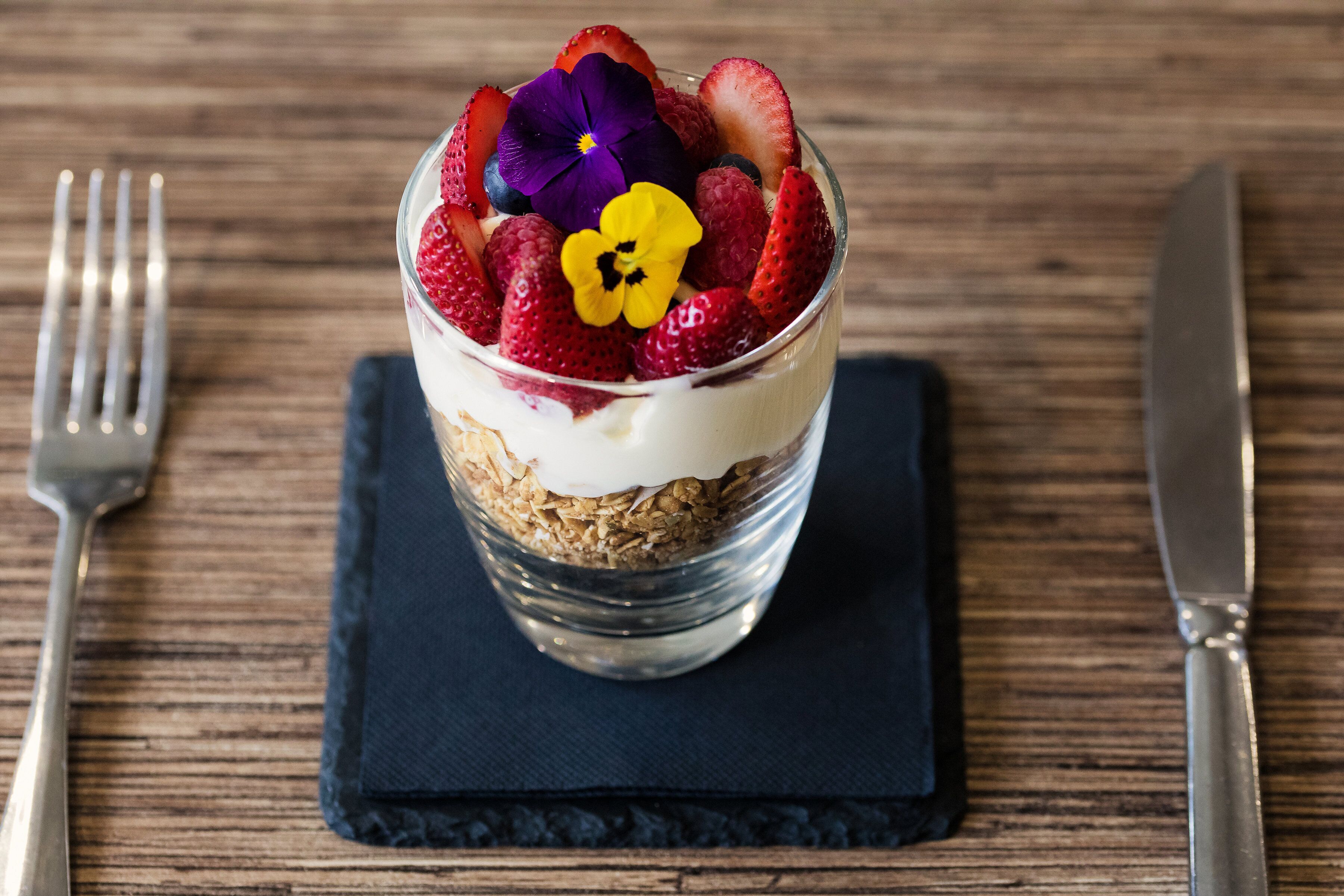 A dessert in a clear glass with strawberries and flowers on top, placed on a dark napkin on a wooden table.