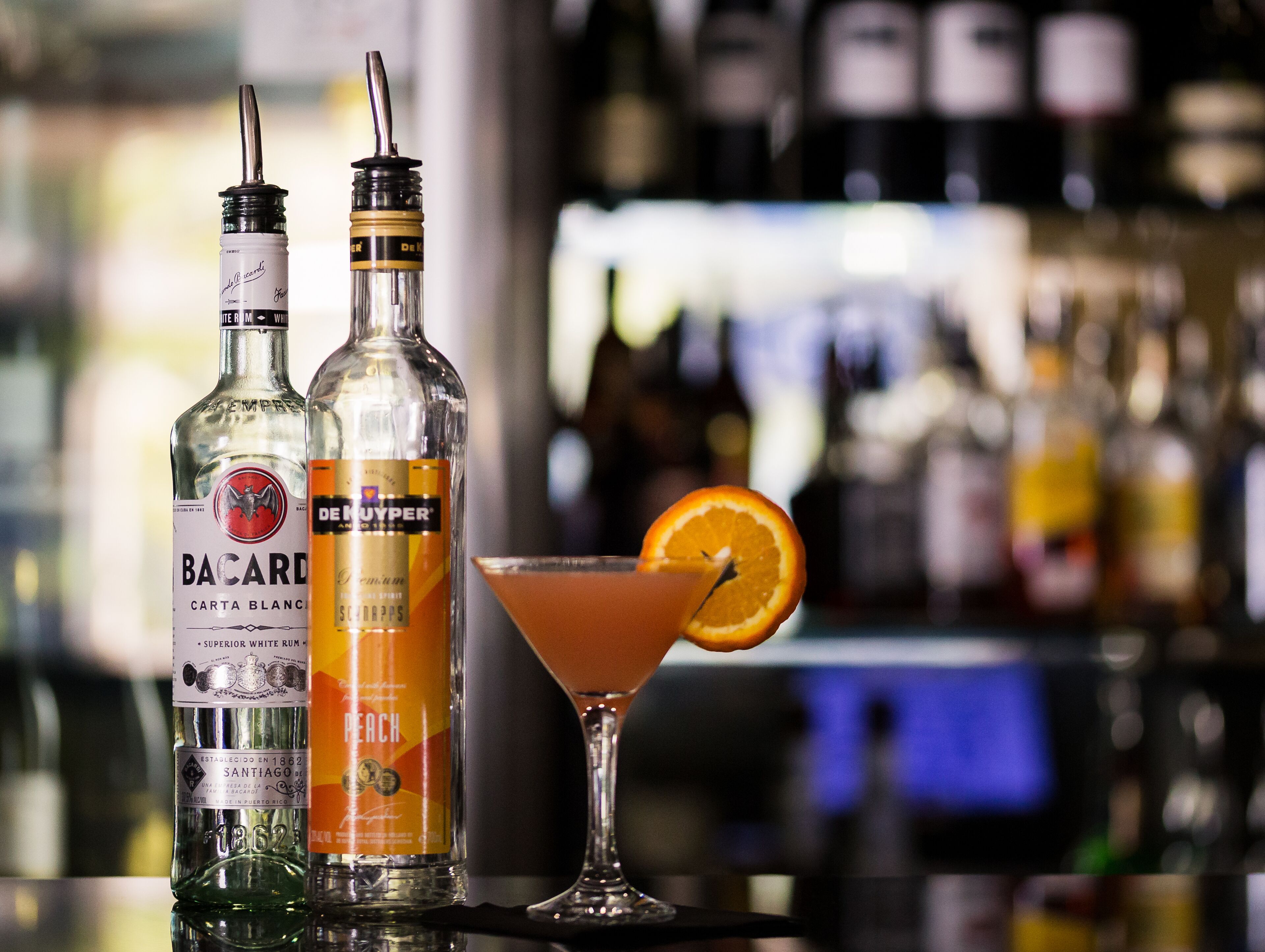 Two bottles of Bacardi and De Nuyper on a bar counter with a cocktail glass containing an orange slice.