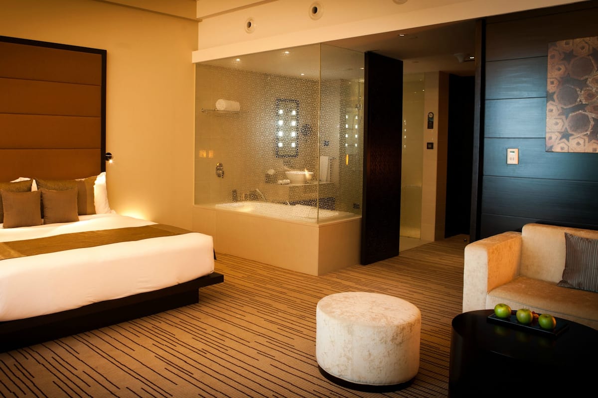A modern hotel room with a large bed, a glass-enclosed bathroom, a seating area, and a small table with apples.