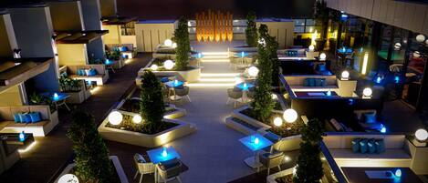 A rooftop lounge with multiple seating areas, including sofas and tables, surrounded by planters and illuminated by ambient lighting.