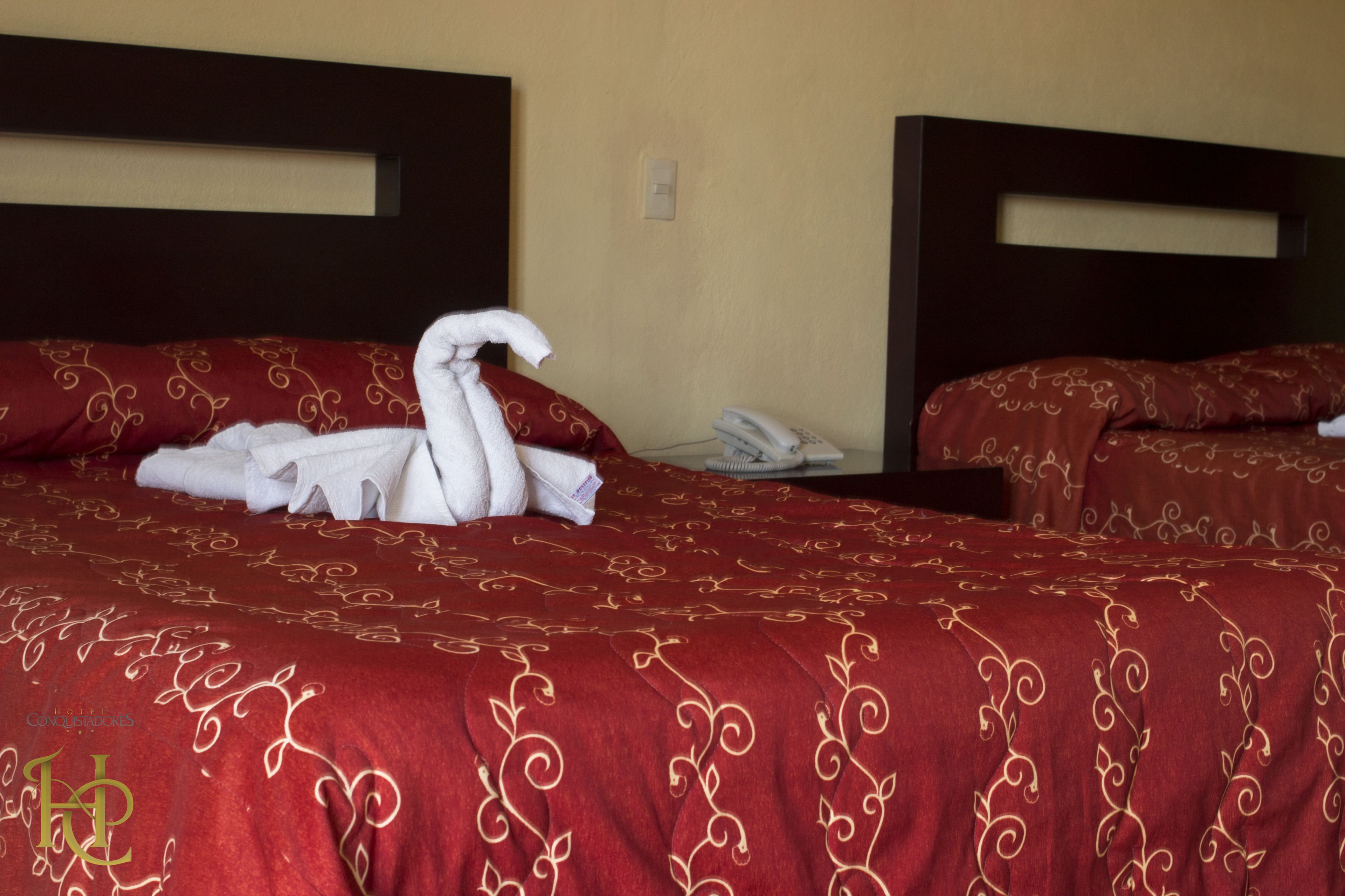 A hotel room with two beds, each with a red bedspread and a dark headboard. There is a white towel arranged in a swan shape on one bed.