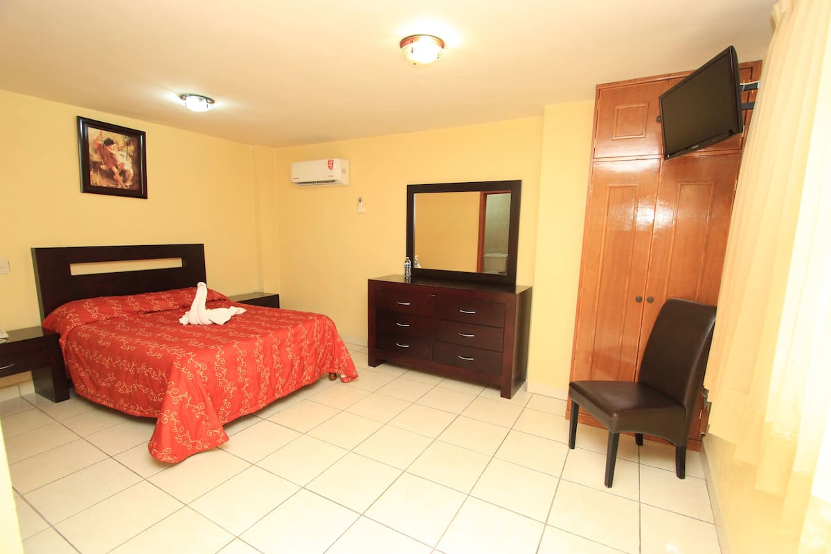 Habitación de hotel con cama, cómoda, silla, televisor montado en la pared y espejo.