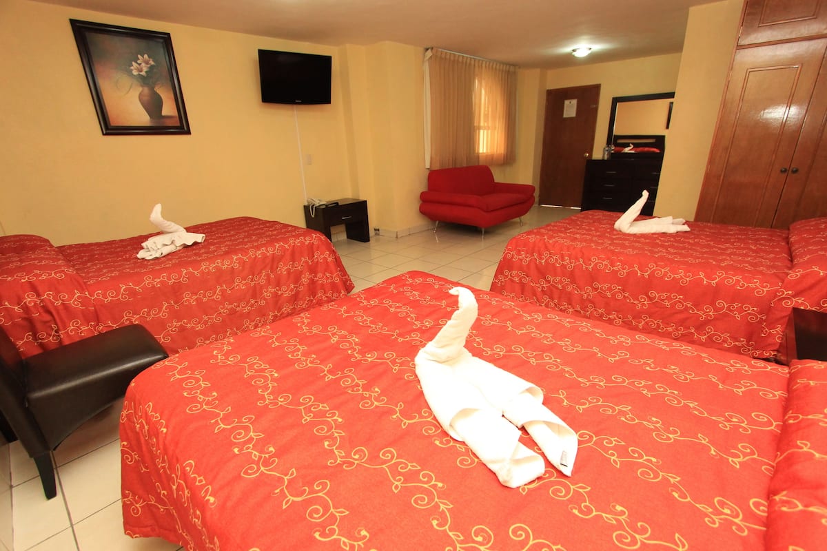 Habitación de hotel con dos camas, cada una con una colcha roja y una toalla blanca doblada encima. Hay un sillón rojo y un armario de madera en la habitación.