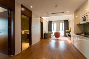 Una habitación de hotel moderna con suelo de madera, un sofá, una silla roja y una zona de cocina con armarios y un microondas.
