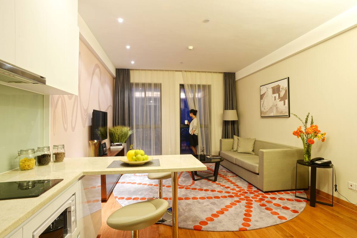 A modern living room with a sofa, a dining table with a bowl of fruit, a kitchen area with an oven, and a large window with curtains.