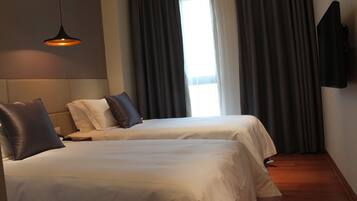 A hotel room with two beds, a flat-screen TV, a large window with curtains, and a hanging pendant light.