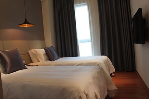 A hotel room with two beds, a flat-screen TV, a large window with curtains, and a hanging pendant light.