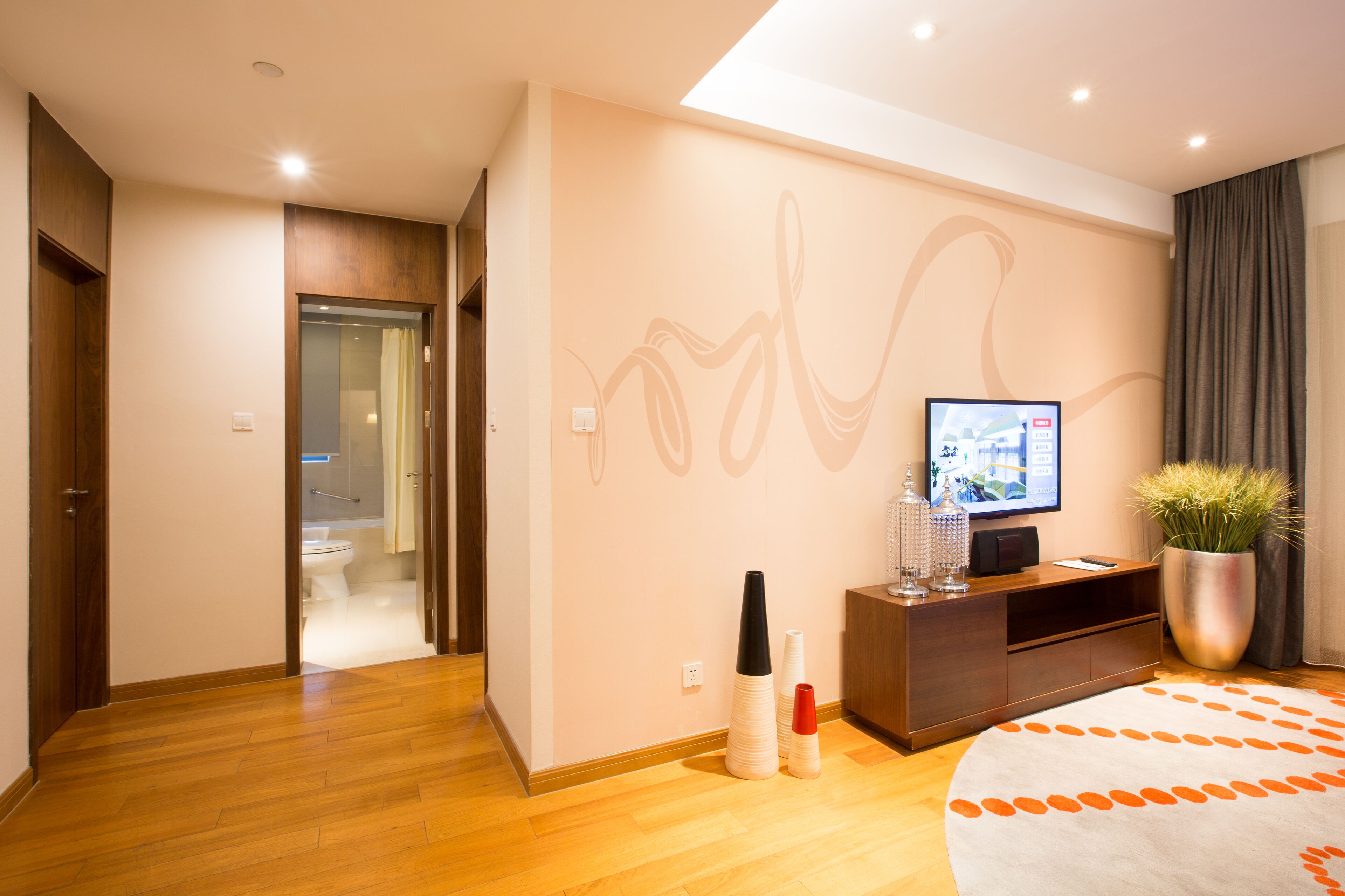 Una habitación de hotel moderna con suelo de madera, una cama grande, un televisor sobre un mueble de madera, una planta en maceta y un baño visible a través de una puerta abierta.