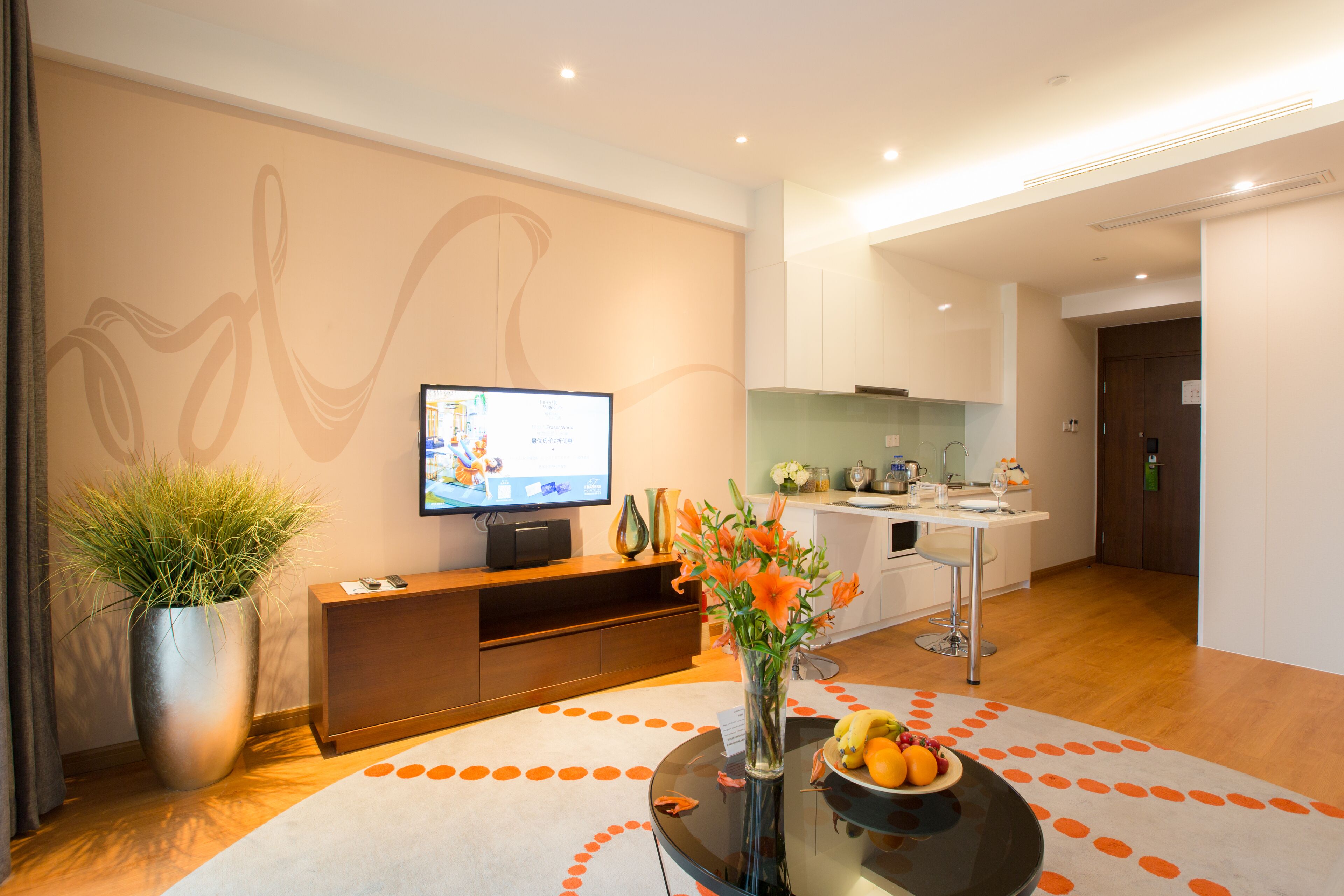 Ein modernes Wohnzimmer mit Flachbildfernseher, einer großen Kübelpflanze, einer Blumenvase und einer Obstschale auf einem Glastisch.