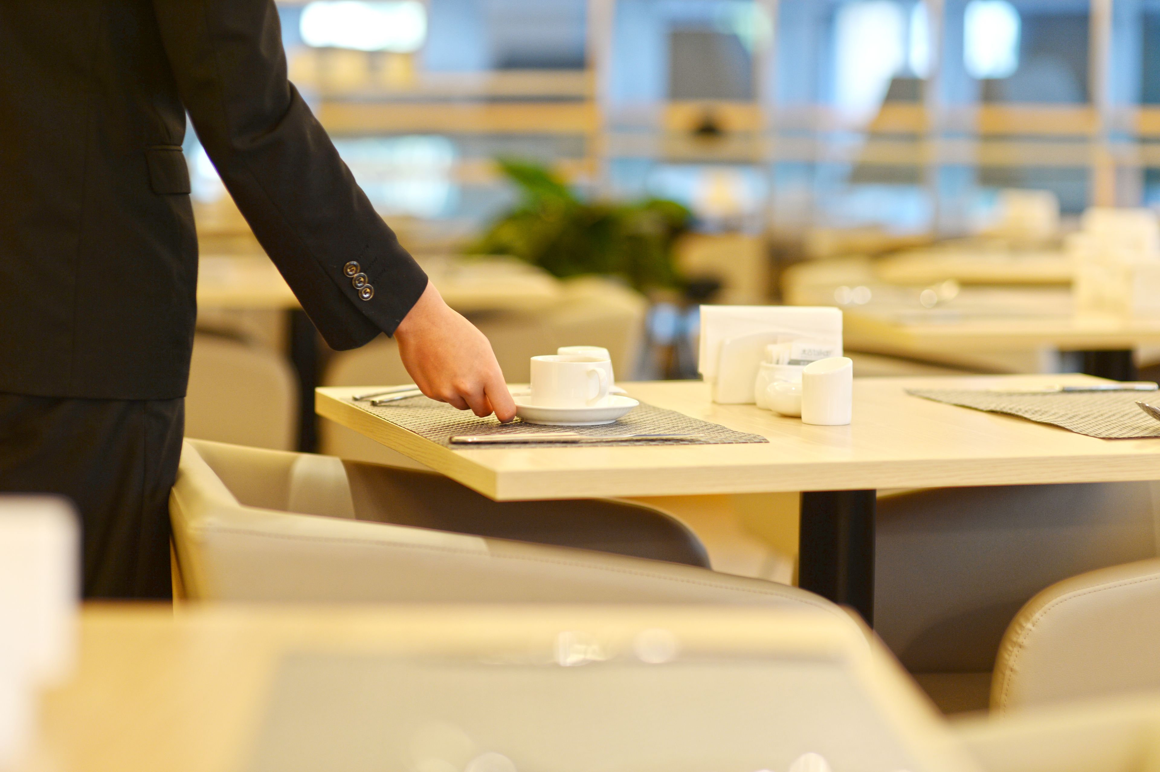 A hotel dining area with tables set for service, including white cups and saucers, and a waiter in a black jacket.