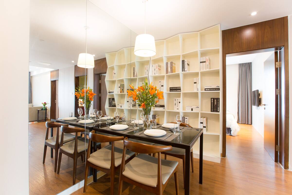 A modern dining area with a glass table, wooden chairs, and a bookshelf in the background.
