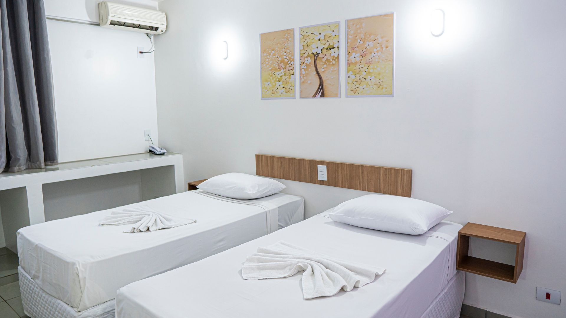 Habitación de hotel con dos camas individuales, cabecero de madera, mesita de noche, aire acondicionado de pared y tres cuadros decorativos.
