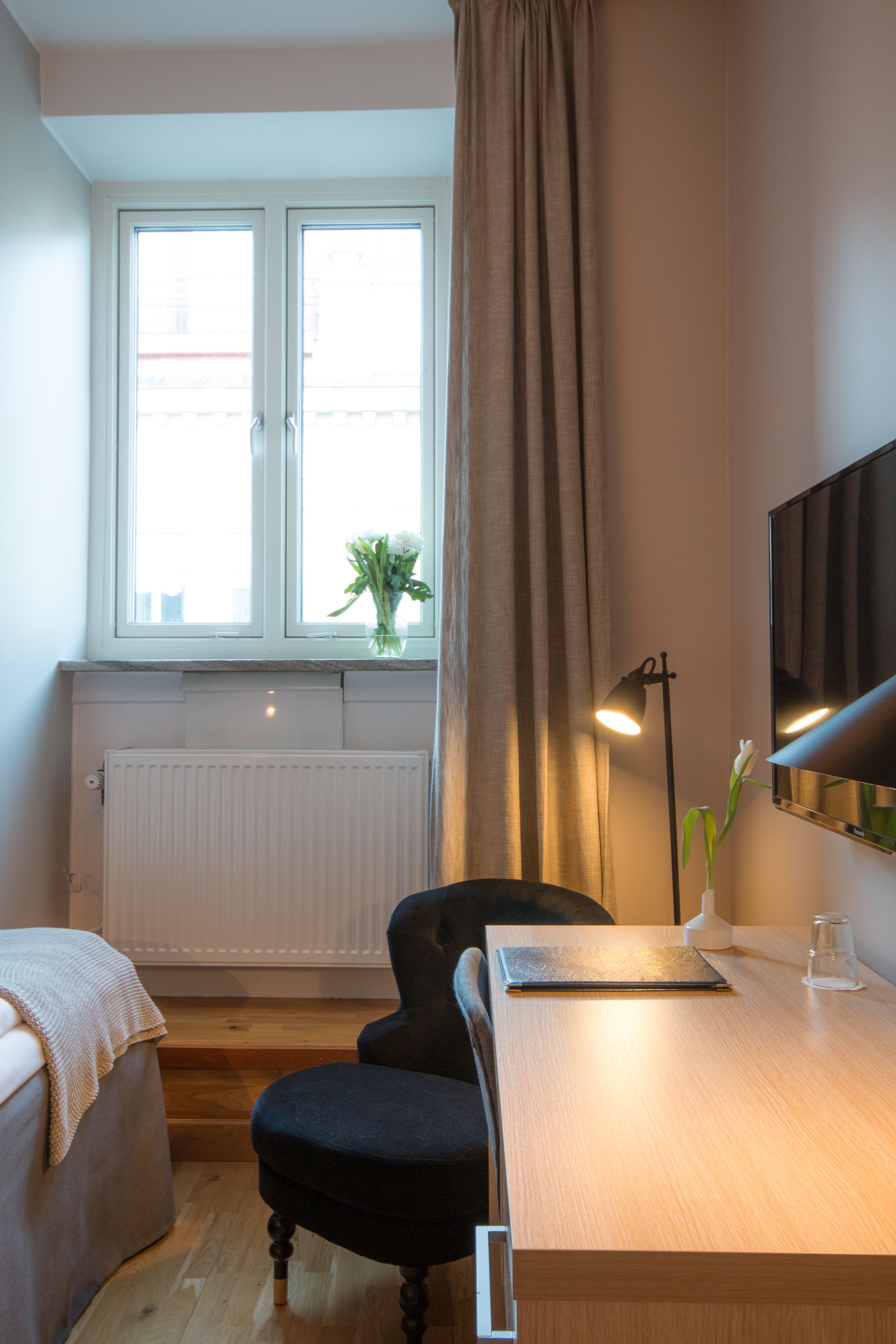 A hotel room with a wooden desk, a chair, a bed, a window with curtains, a radiator, and a wall-mounted TV.