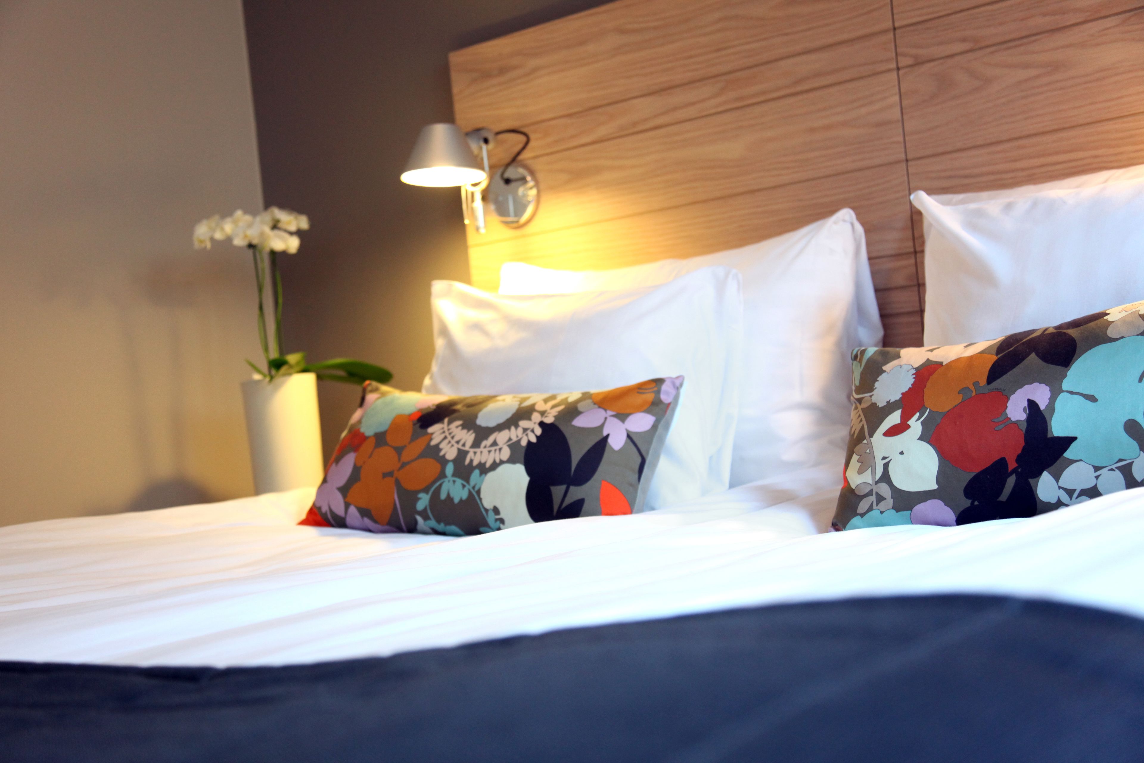 Una cama bien hecha con sábanas blancas y almohadillas florales de colores, un cabecero de madera y una lámpara fijada a la pared.