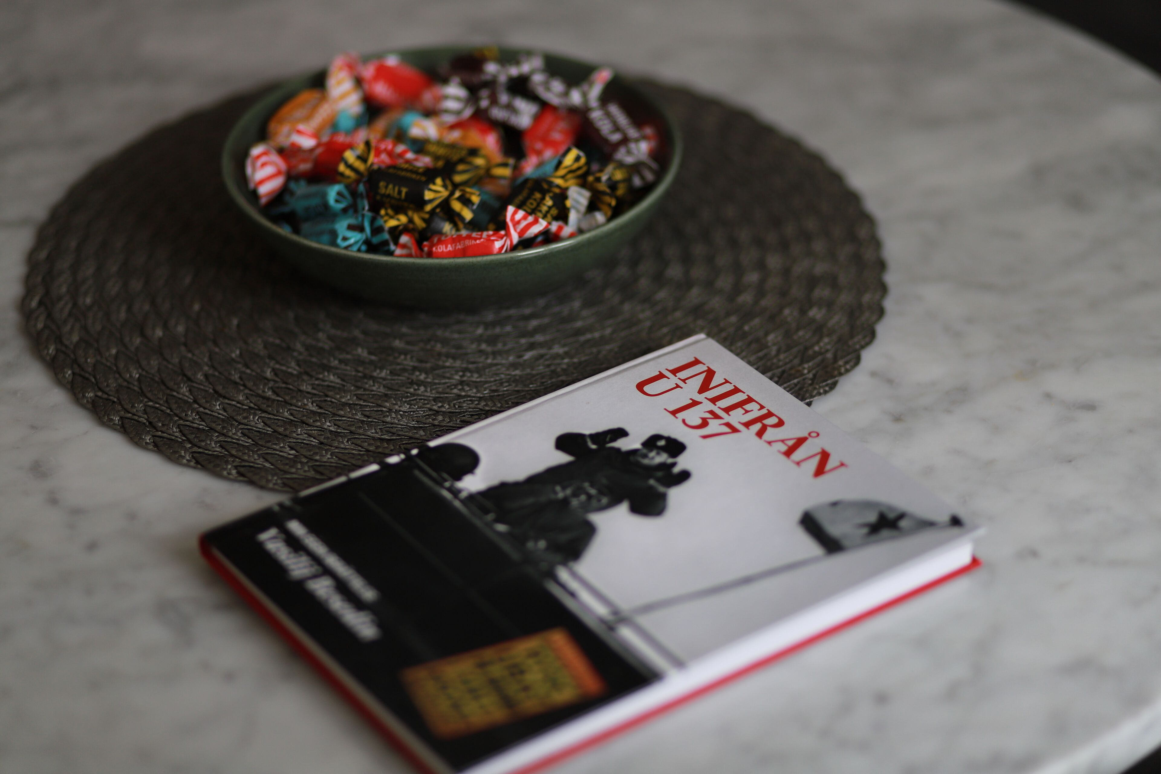 A book titled "INFERNO 137" and a bowl of assorted candies on a woven placemat.