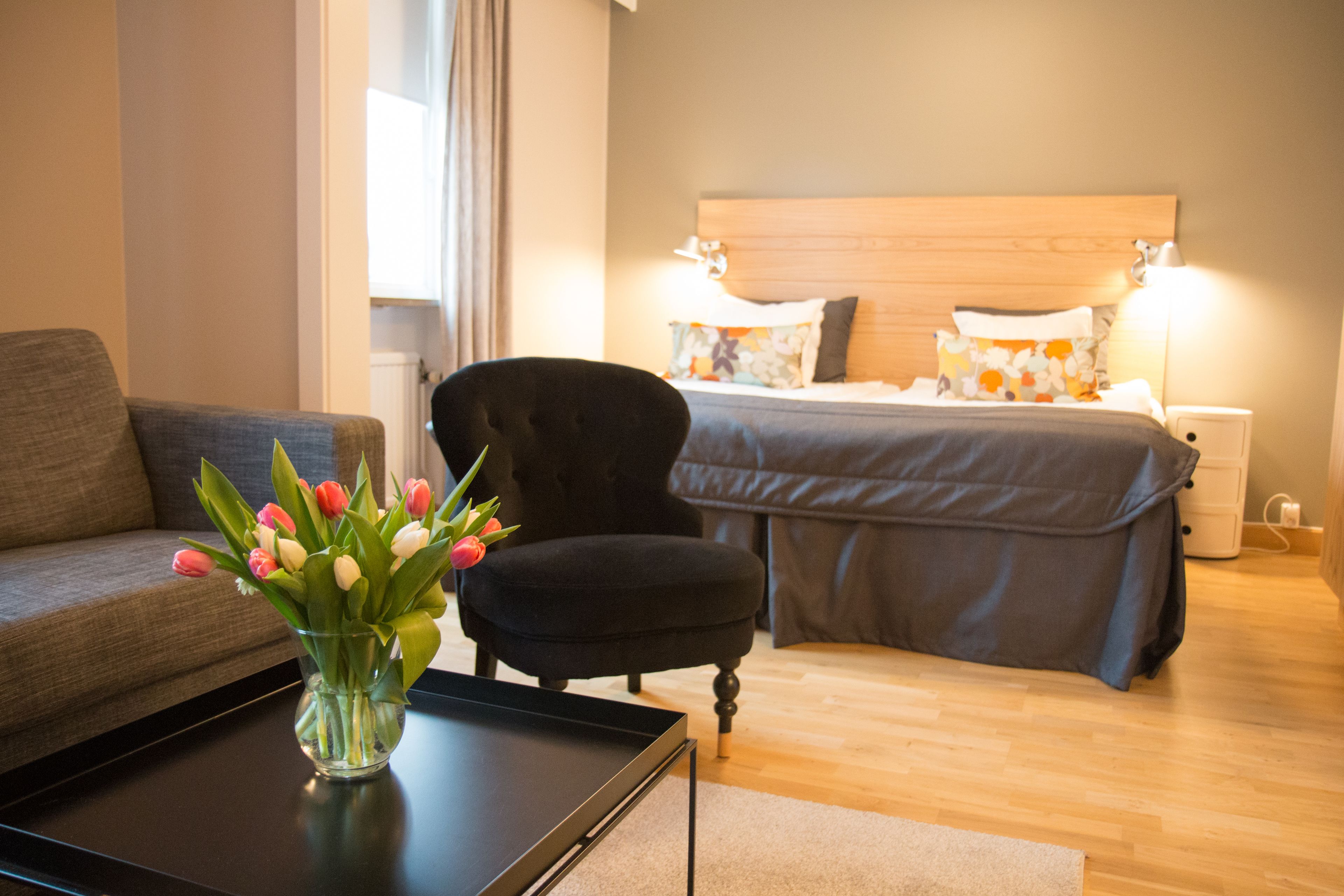 Quarto de hotel com cama, sofá, cadeira e mesa de centro com um vaso de flores.