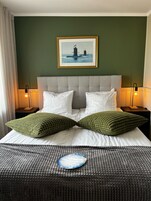 Una habitación de hotel con una cama grande, dos lámparas de noche, un cuadro con faros y un cojín decorativo.