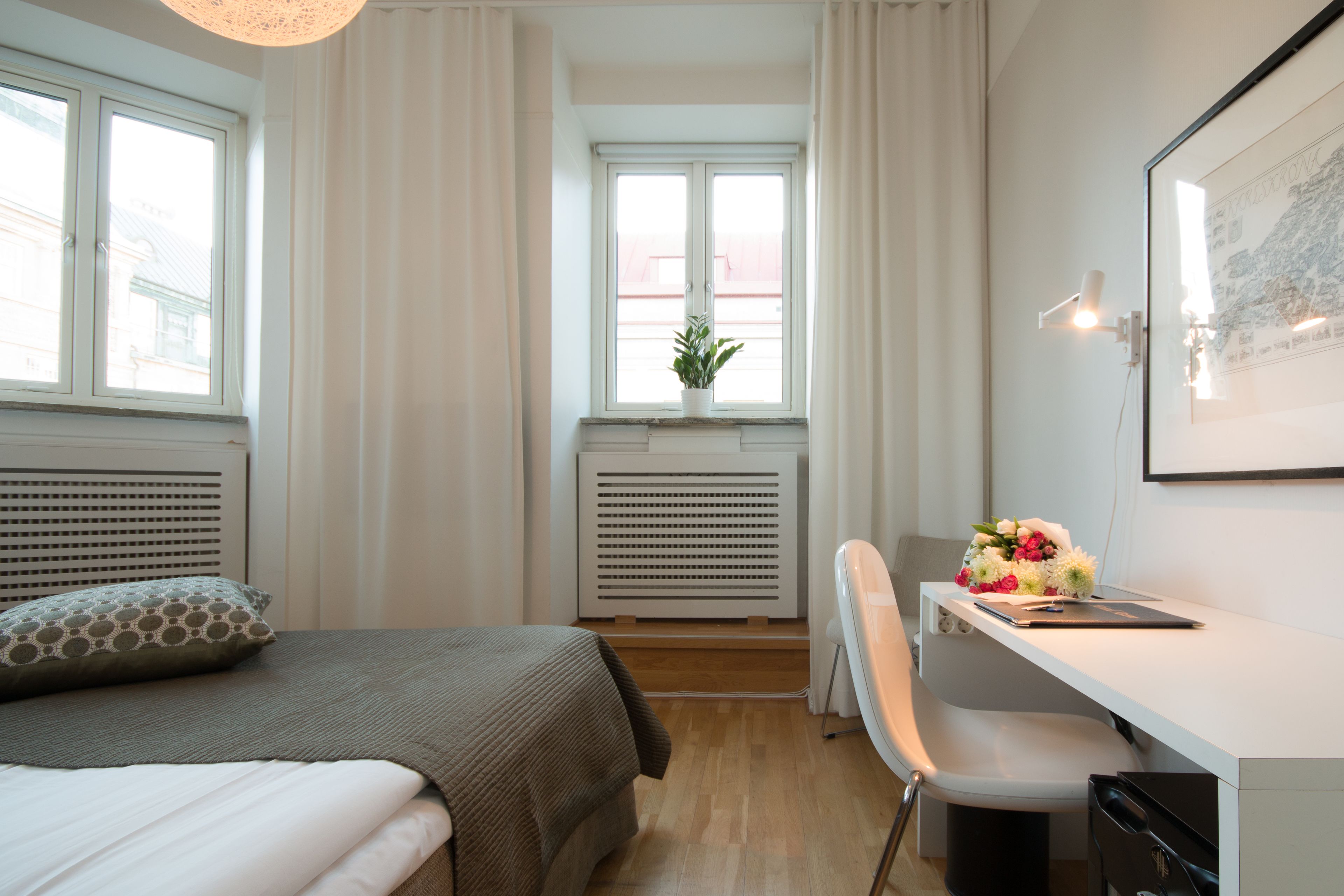 Une chambre à coucher avec un lit, un bureau, une chaise, un radiateur et une fenêtre avec des rideaux.